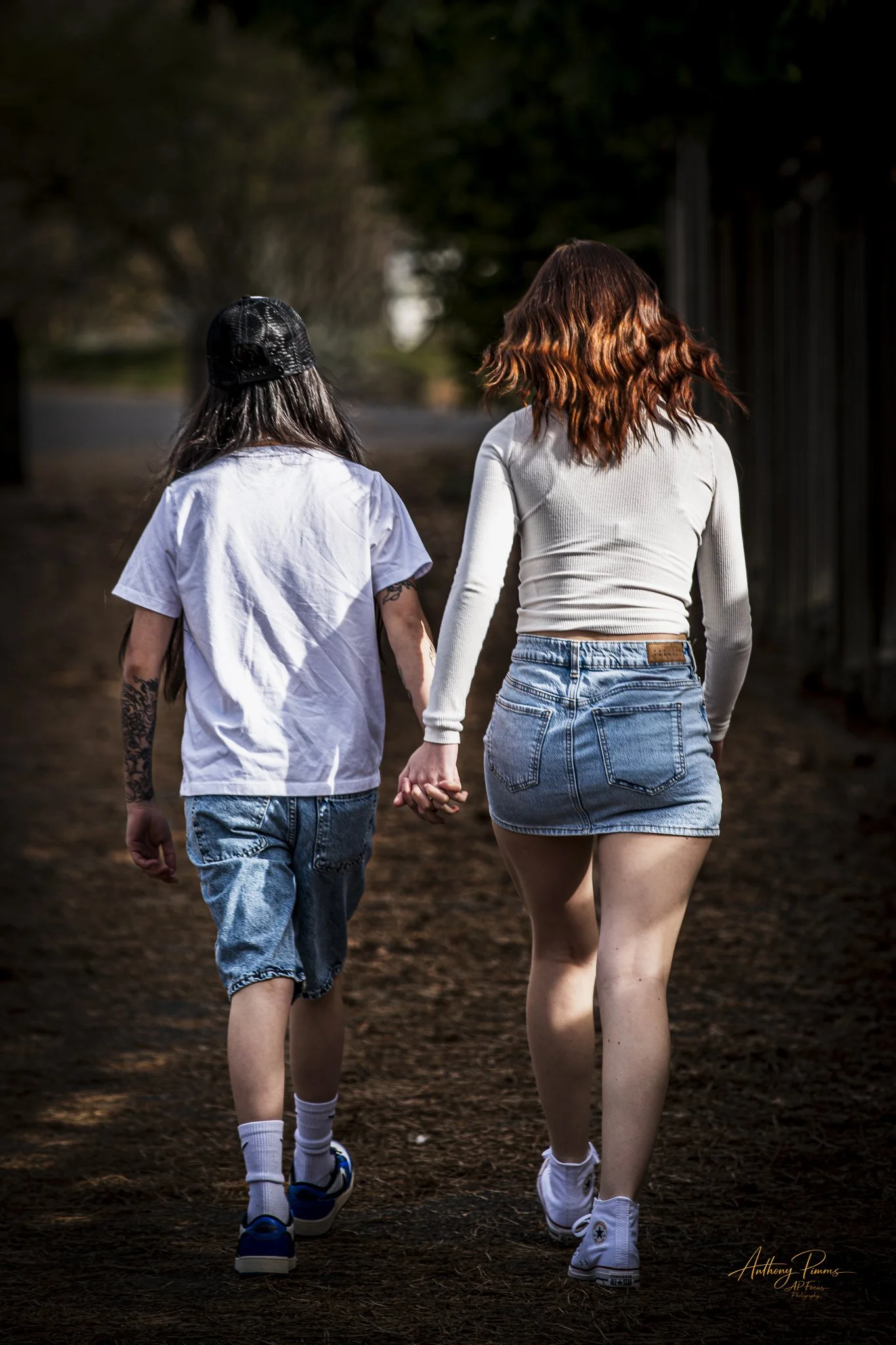 Two young women holding hands and walking away on a dark wooded trail, with long hair and casual clothing, during daylight.