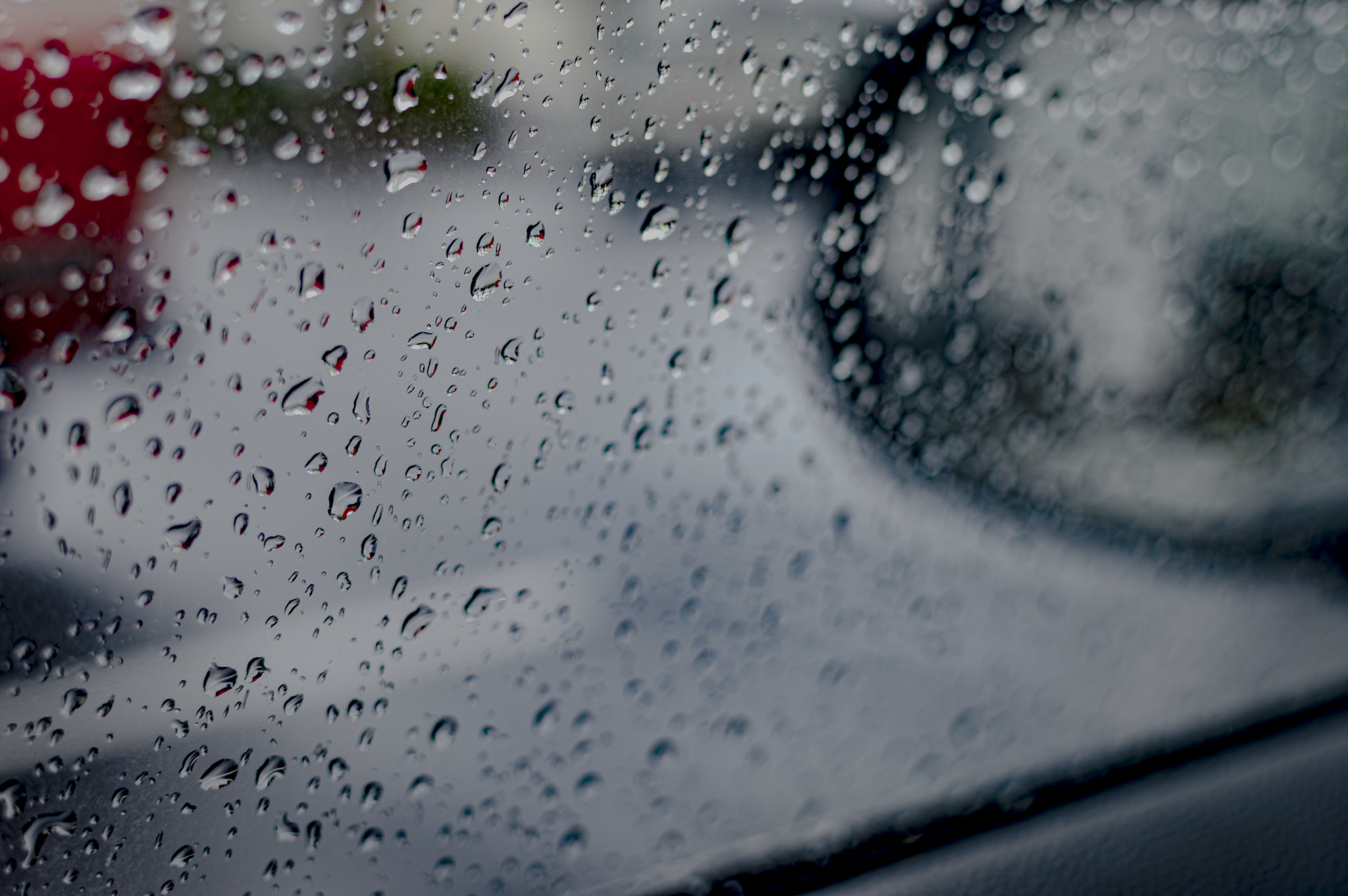 Raindrops on a car window with a blurry view of the outside, including a red object and cloudy sky in the background.