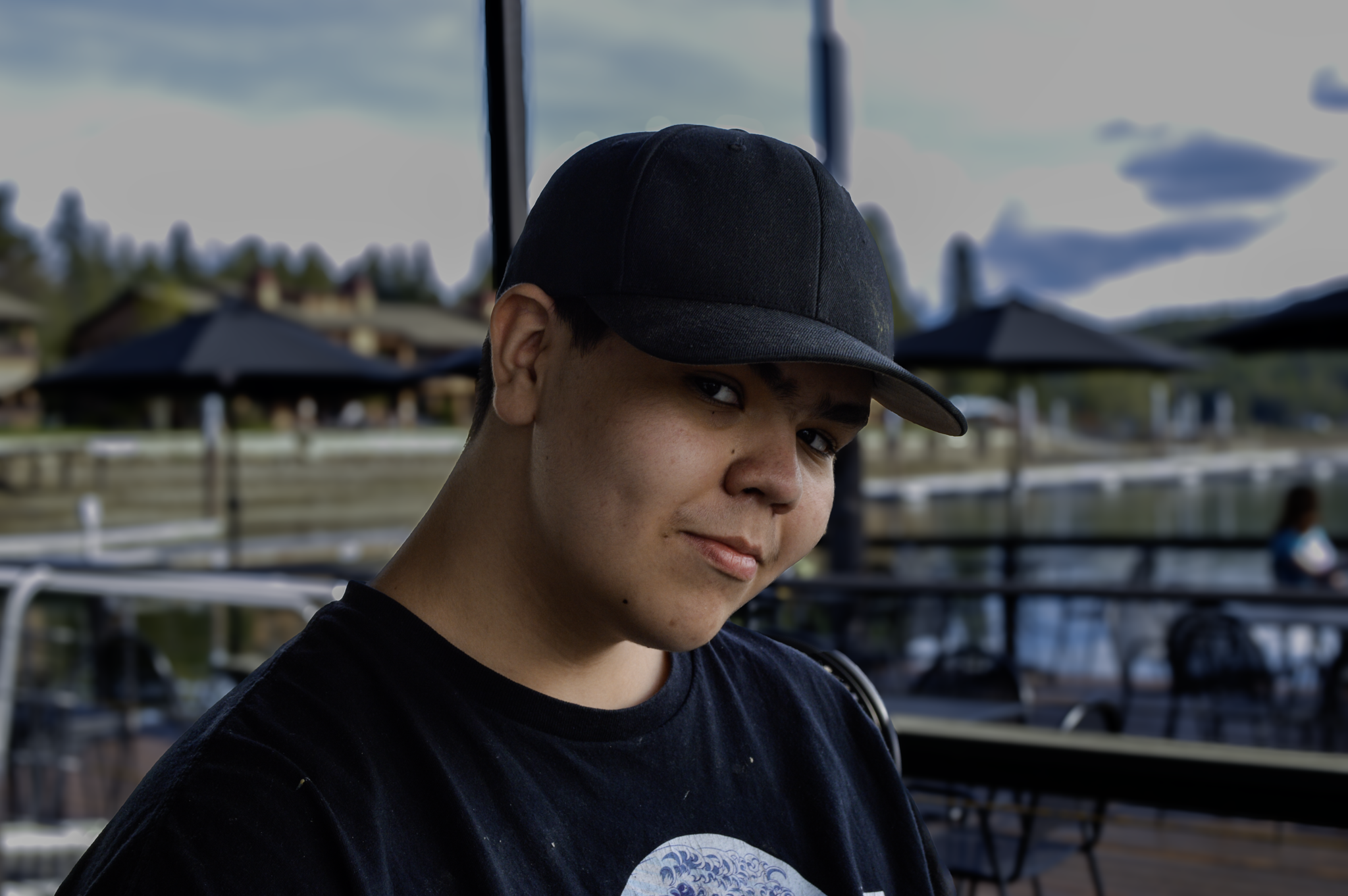 A close-up of a young person's face, wearing a black cap and black shirt, with an outdoor background including umbrellas and a wooden deck.