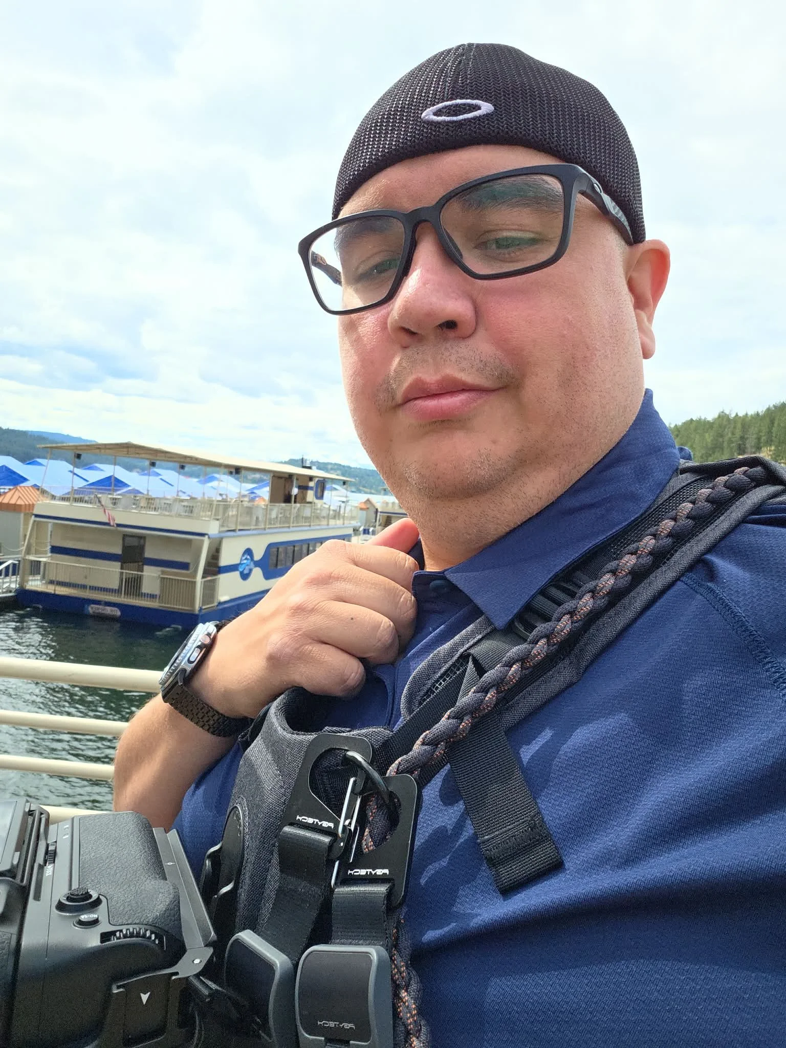 A man wearing glasses and a black beanie hat with an oval logo, carrying a backpack, holding a camera, and standing near a body of water with a dock and boats in the background.