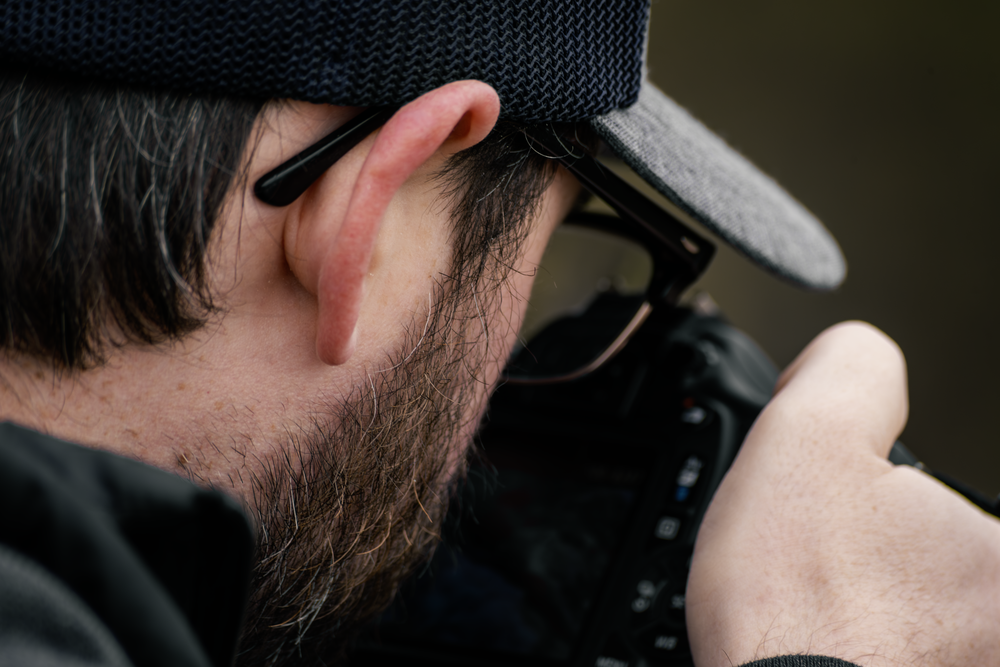 A close-up of a man wearing a gray and white cap, black glasses, and a black shirt, holding and looking at a smartphone, with a beard and brown hair visible.