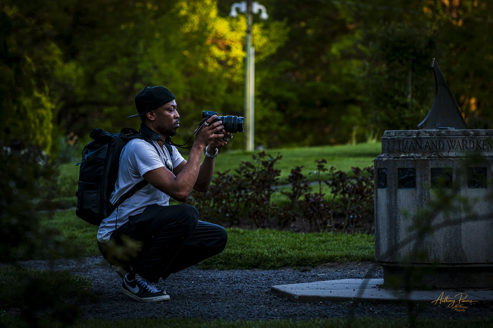A man wearing a black cap, white T-shirt, and a backpack is crouched down in a park during daylight, holding a camera and taking a photograph of a memorial plaque on a stone monument surrounded by greenery.