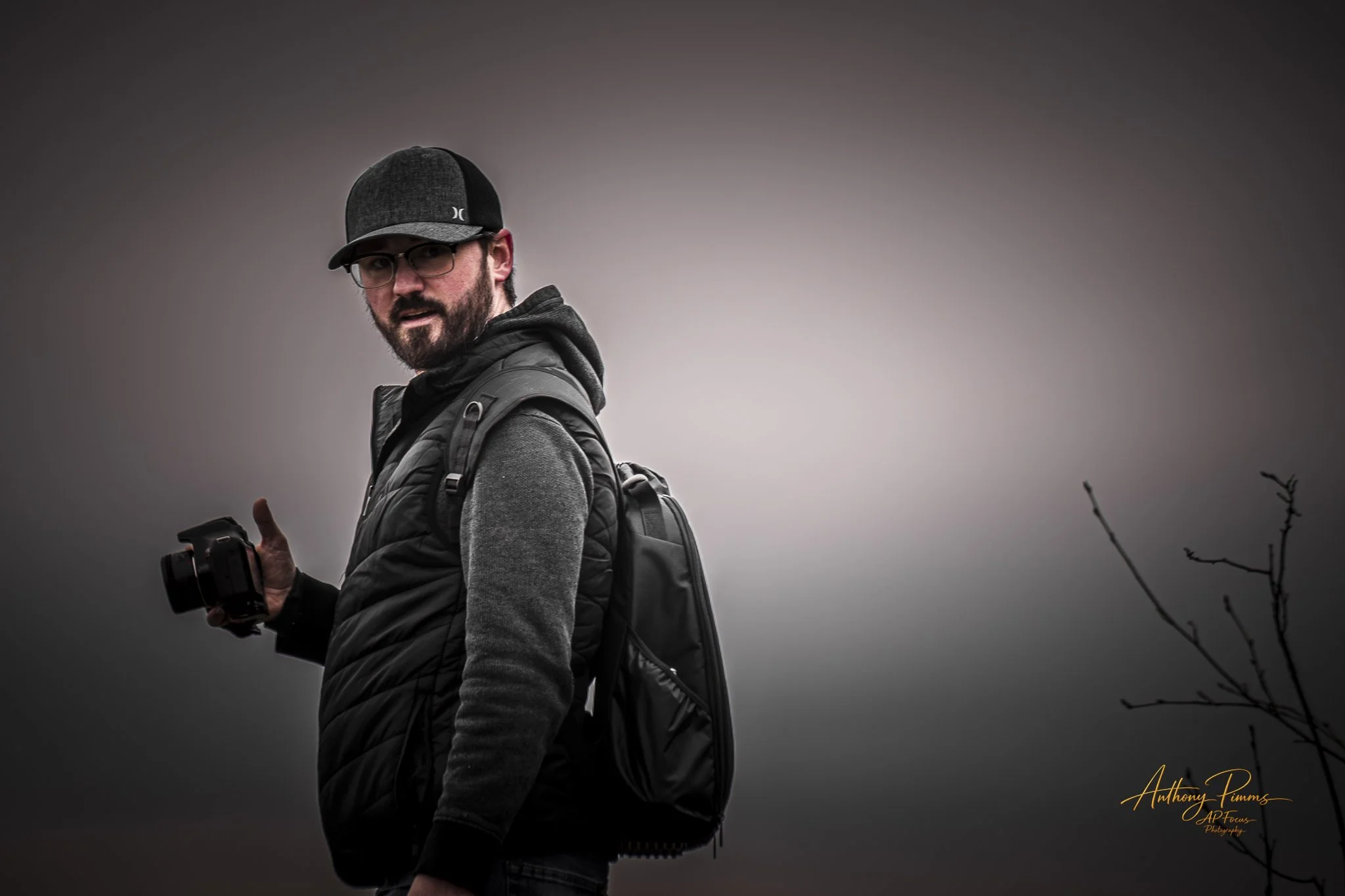 A man with glasses, a beard, and a hat, wearing a gray jacket and backpack, holding a camera, standing outdoors against a cloudy sky with some branches in the foreground.