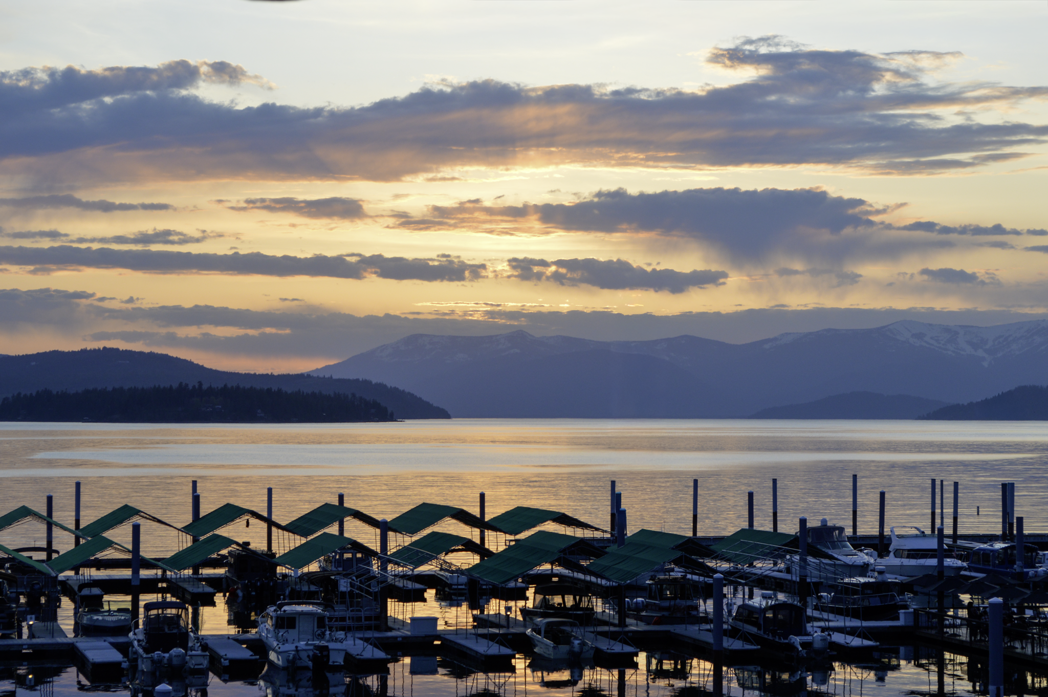 A marina with boats docked under green awnings, tranquil water reflecting the sky, mountains in the background, and a partly cloudy sunset sky.