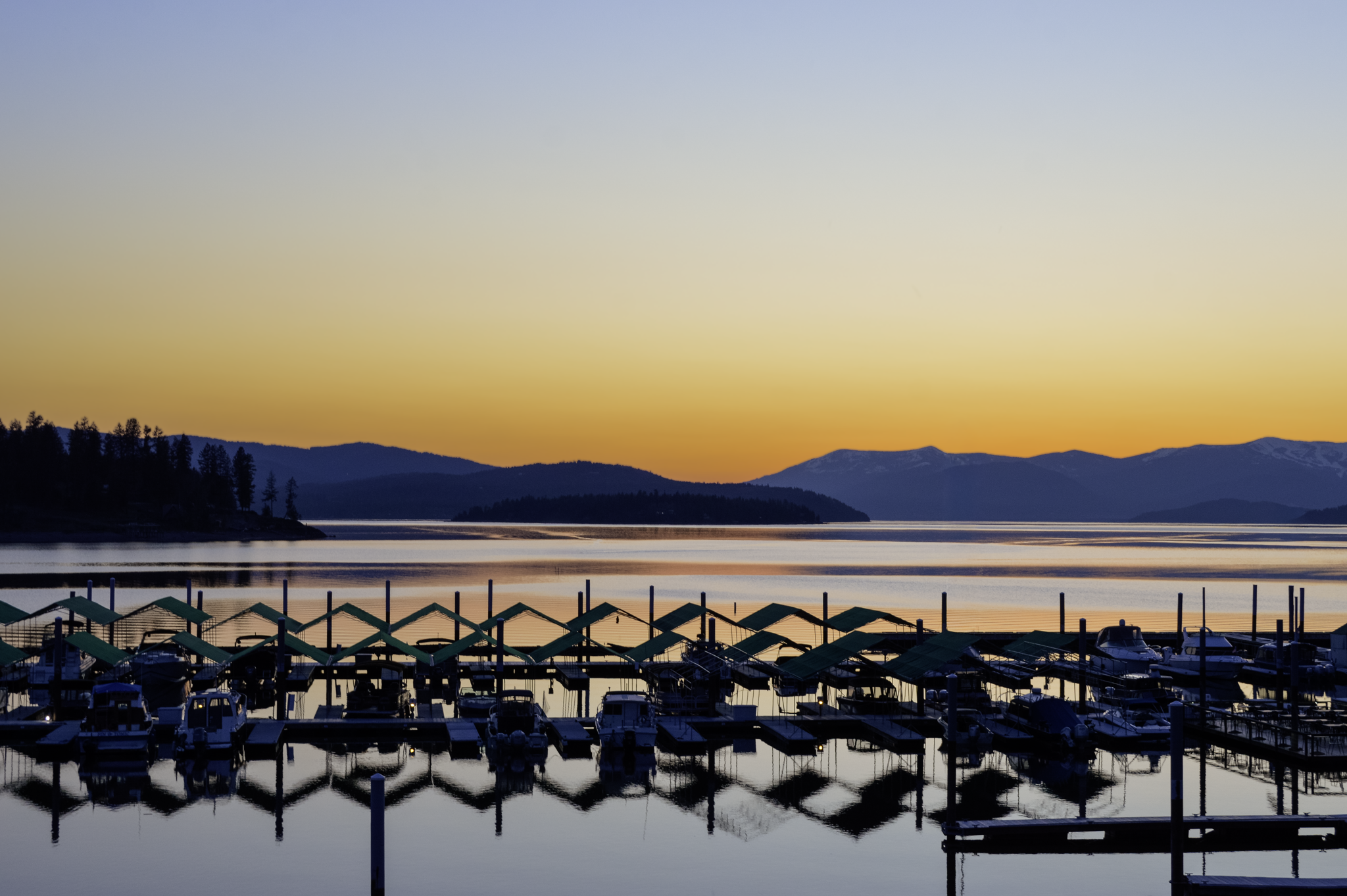 View of a marina at sunset with boats docked under green canopies, calm water reflecting the surroundings, distant mountains, and a clear sky fading from yellow to blue.