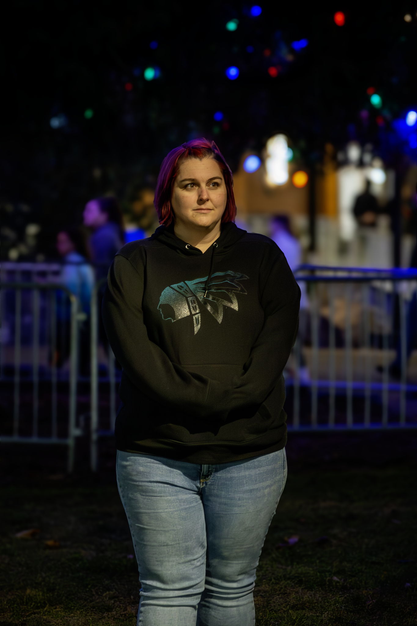 Young woman with purple hair wearing a black hoodie and light-colored jeans at night with blurred colorful lights in the background.