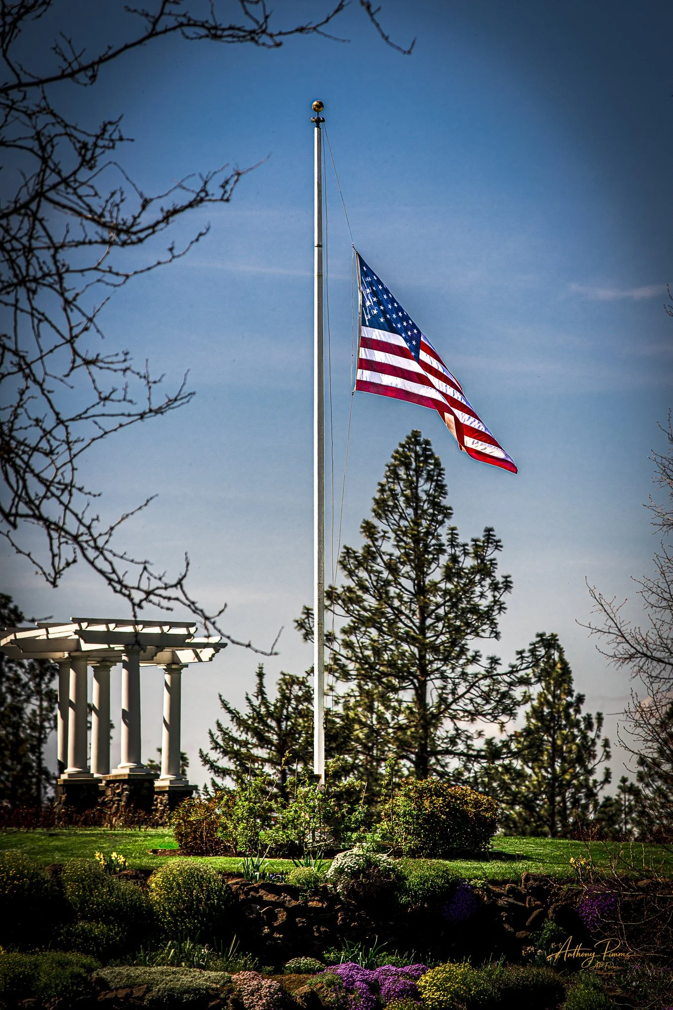 American flag flying on a flagpole in a landscaped garden, with a white classical temple structure and trees in the background during daytime.