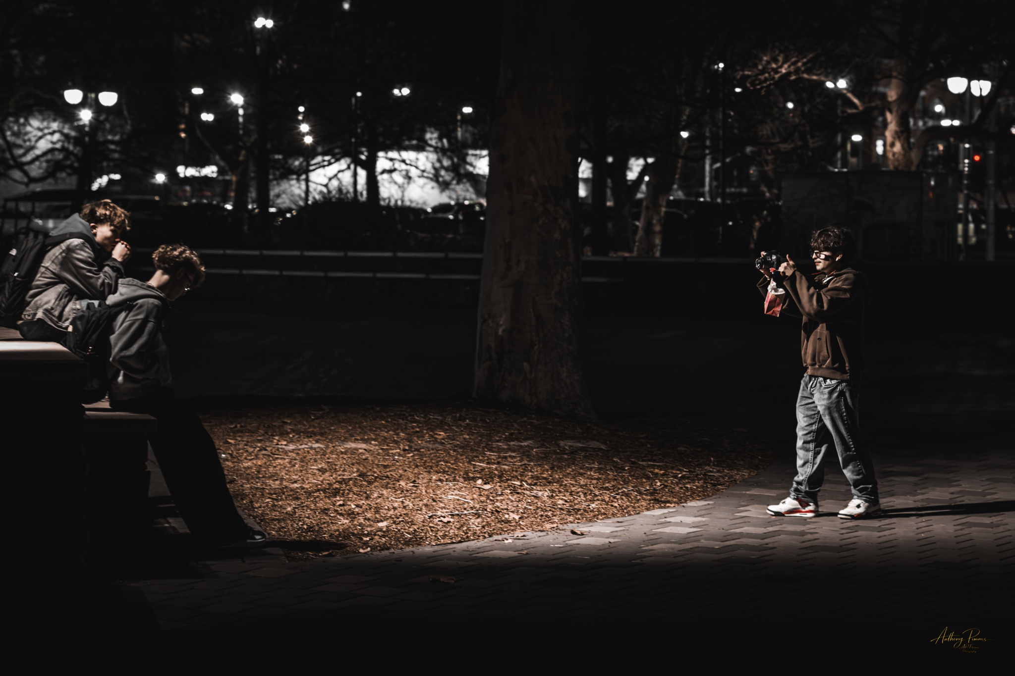 A nighttime scene in a park with trees and streetlights. Two people are sitting on a bench, whispering or talking, and a young boy taking a photograph with a camera. The ground has fallen leaves, and a large tree trunk is in the center. The backgroun