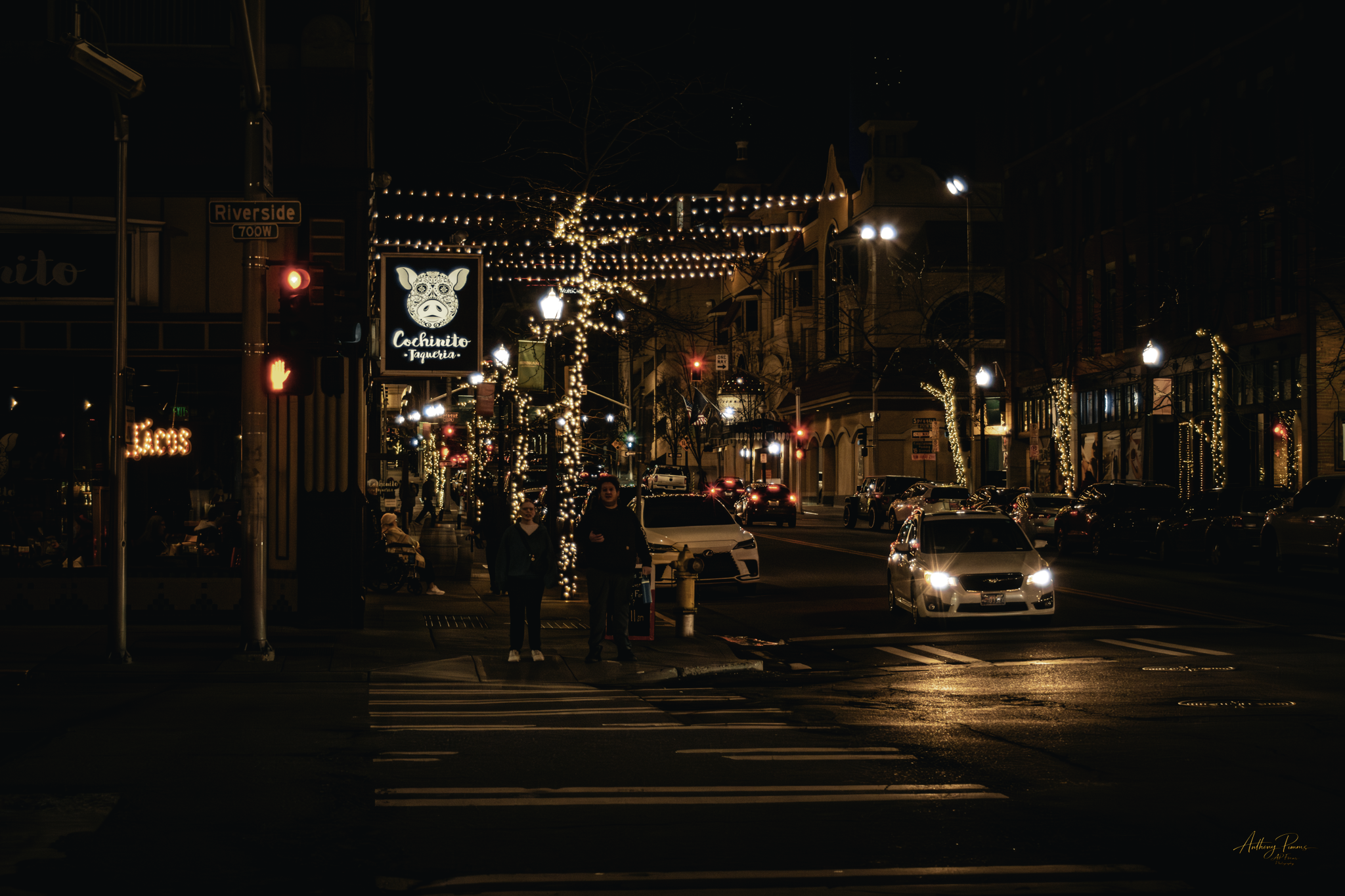 Night street scene with streetlights, decorative string lights, a sign for Cochinito Taqueria, and cars on the road. Pedestrians wait at crosswalks.