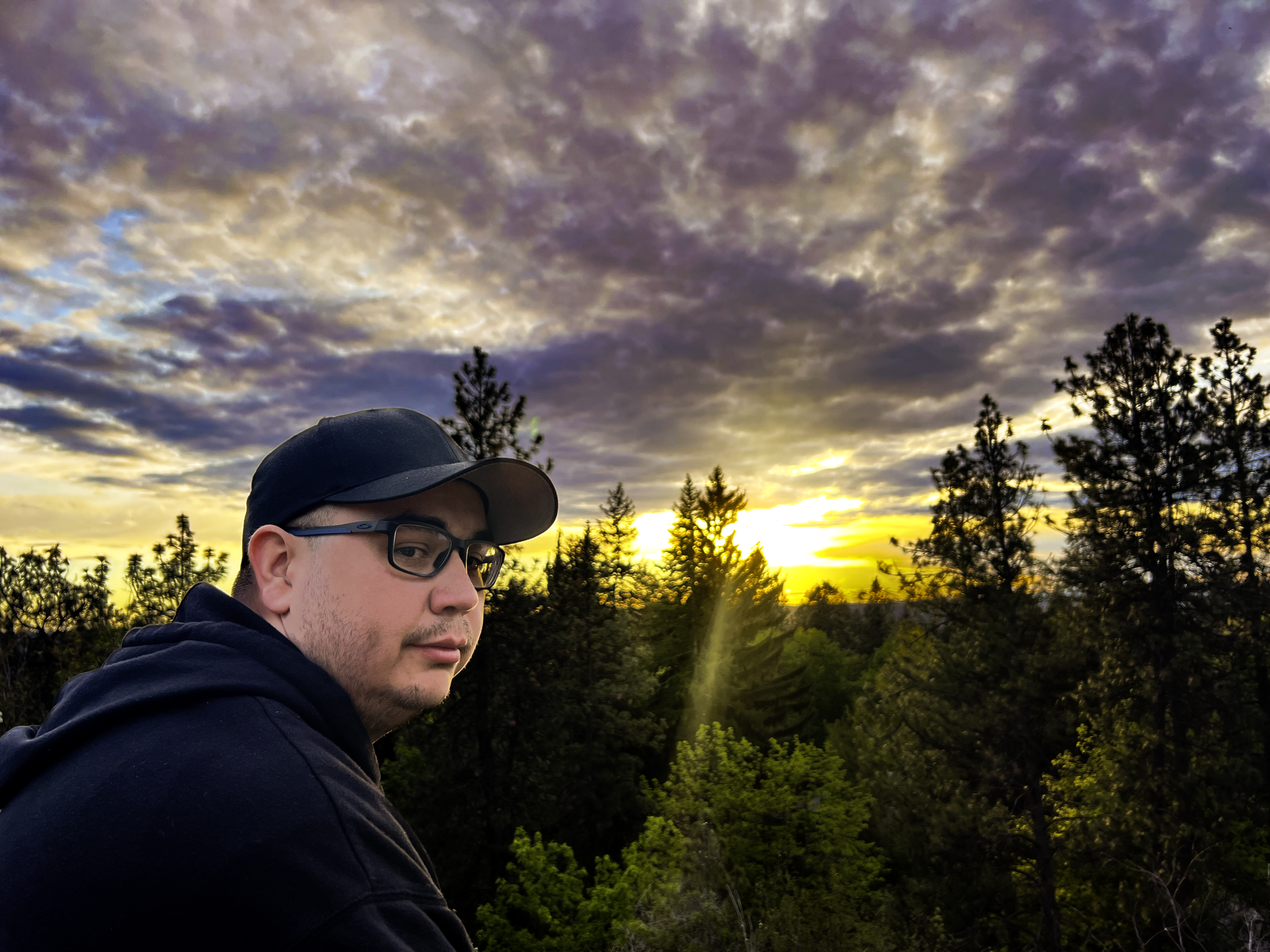 A man wearing glasses and a black baseball cap stands outdoors during sunset with a cloudy sky and trees in the background.