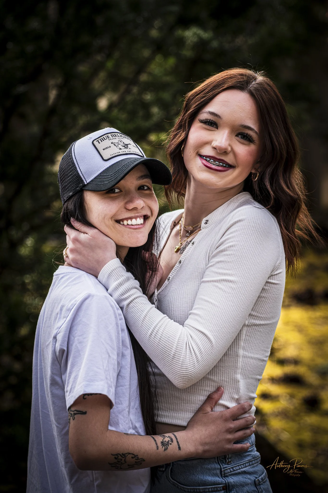 Two women standing outdoors, embracing and smiling at the camera. One woman has long dark hair and is wearing a white shirt and a gray cap with a patch. The other has auburn hair, braces, and is wearing a beige top. In the background, there are trees