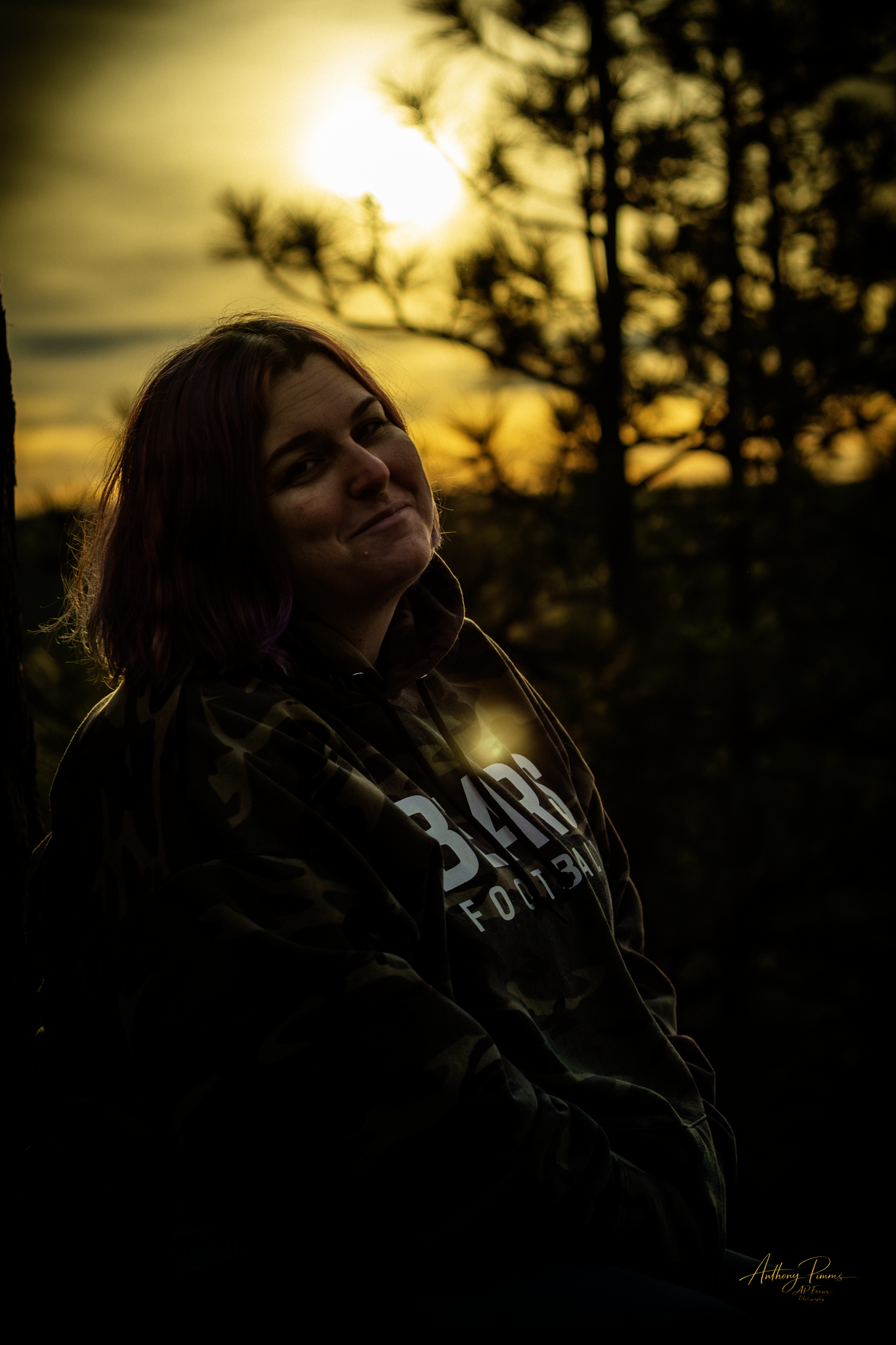 A woman with shoulder-length hair smiling at the camera in a camo hoodie, standing outdoors during sunset with trees and the sun in the background.