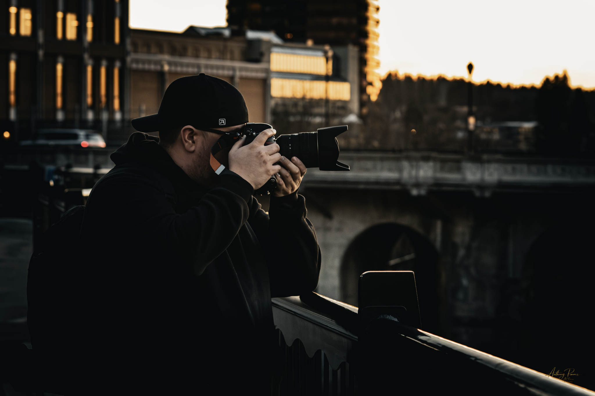 A person wearing a black hoodie and black cap holding a professional camera, taking a photo during sunset near a river or canal with buildings and trees in the background.
