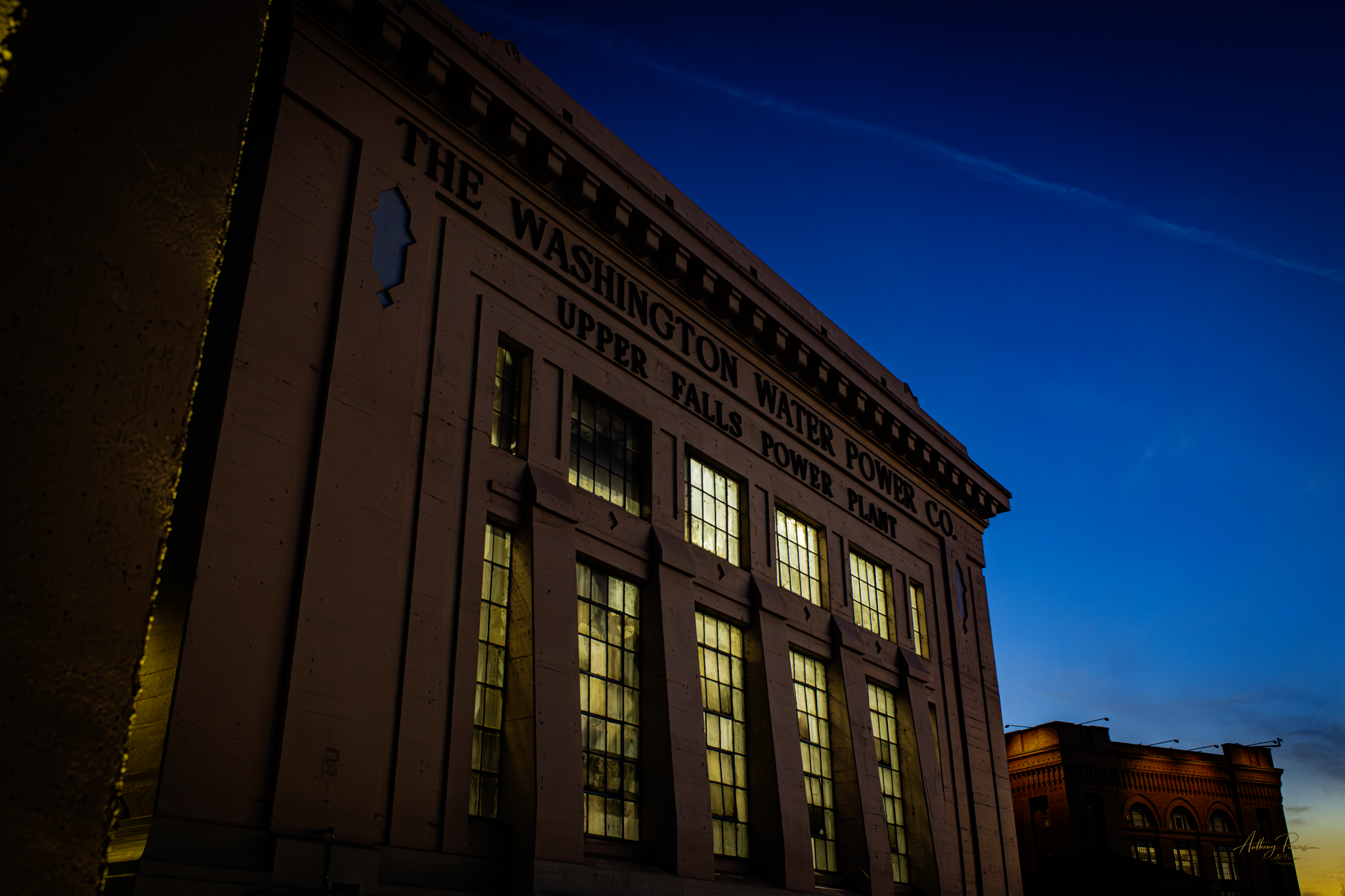A historic building at twilight with illuminated windows and a rooftop sign that reads 'THE WASHINGTON WATER POWER CO.' The sky is dark blue with streaks of light clouds.