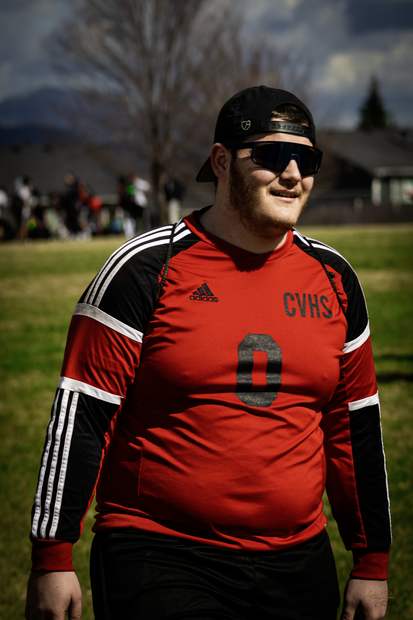 A young man with a beard smiling, wearing a black cap backwards, dark sunglasses, a red and black sports jersey with white stripes on the sleeves, and black shorts, standing outdoors on a sunny day with a grassy area and trees in the background.