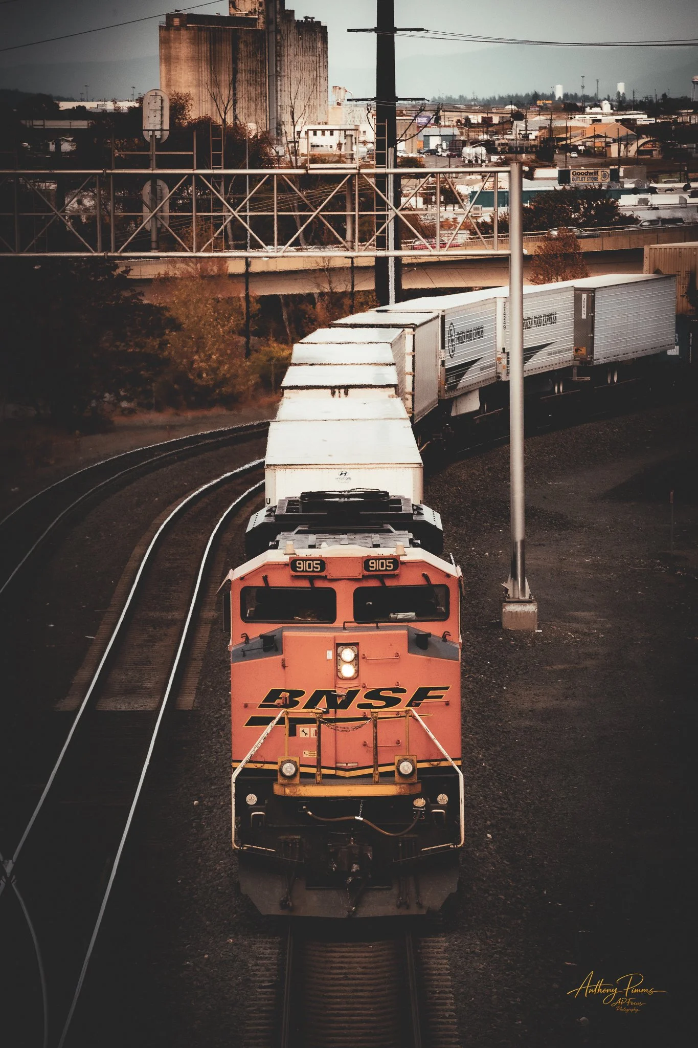 A BNSF freight train moving along curved railroad tracks in an industrial area, with a cityscape background featuring various buildings and industrial structures.
