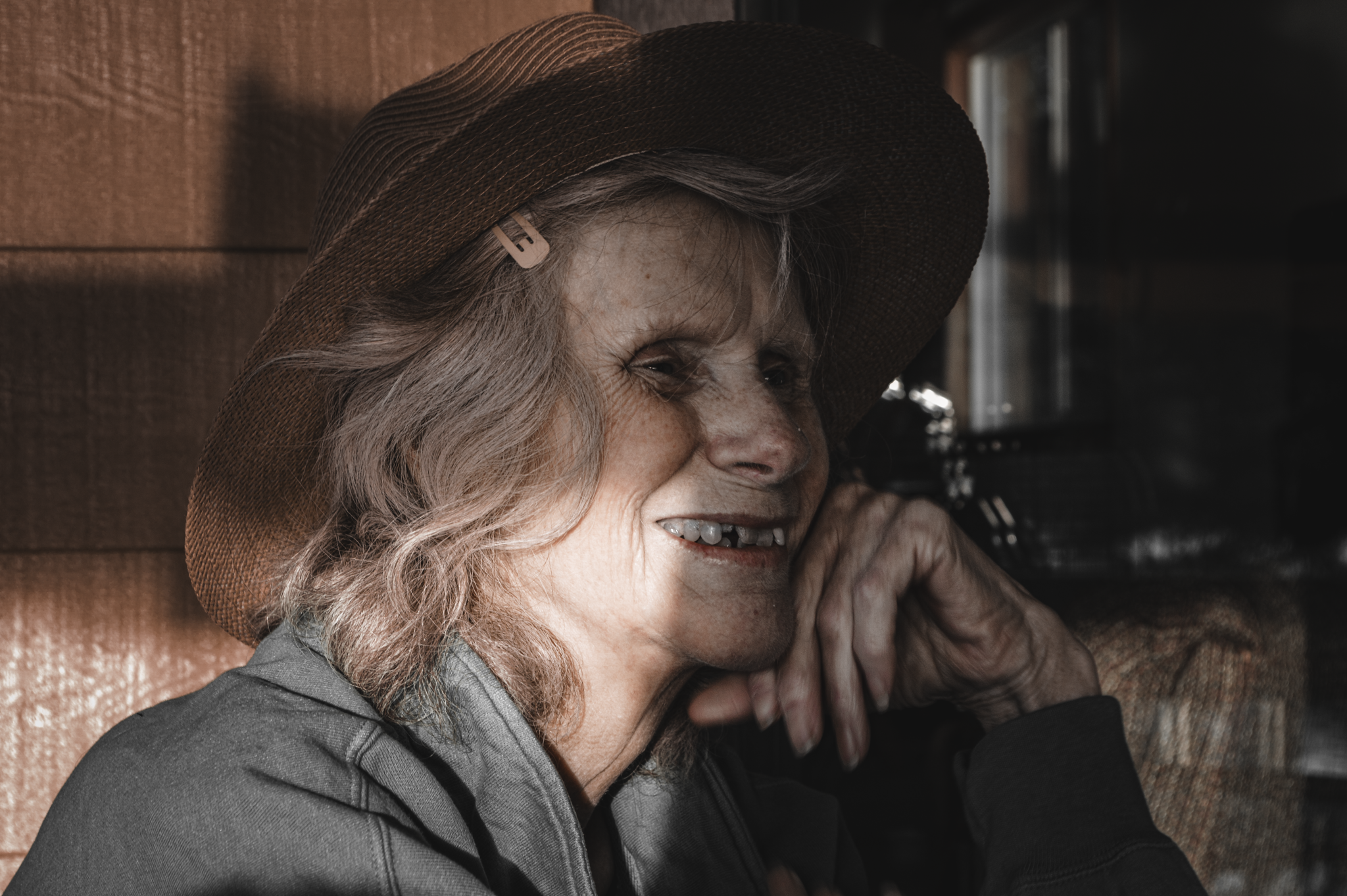 Close-up of an elderly woman with gray hair, wearing a large brown hat with a hair clip, smiling and leaning on her hand.