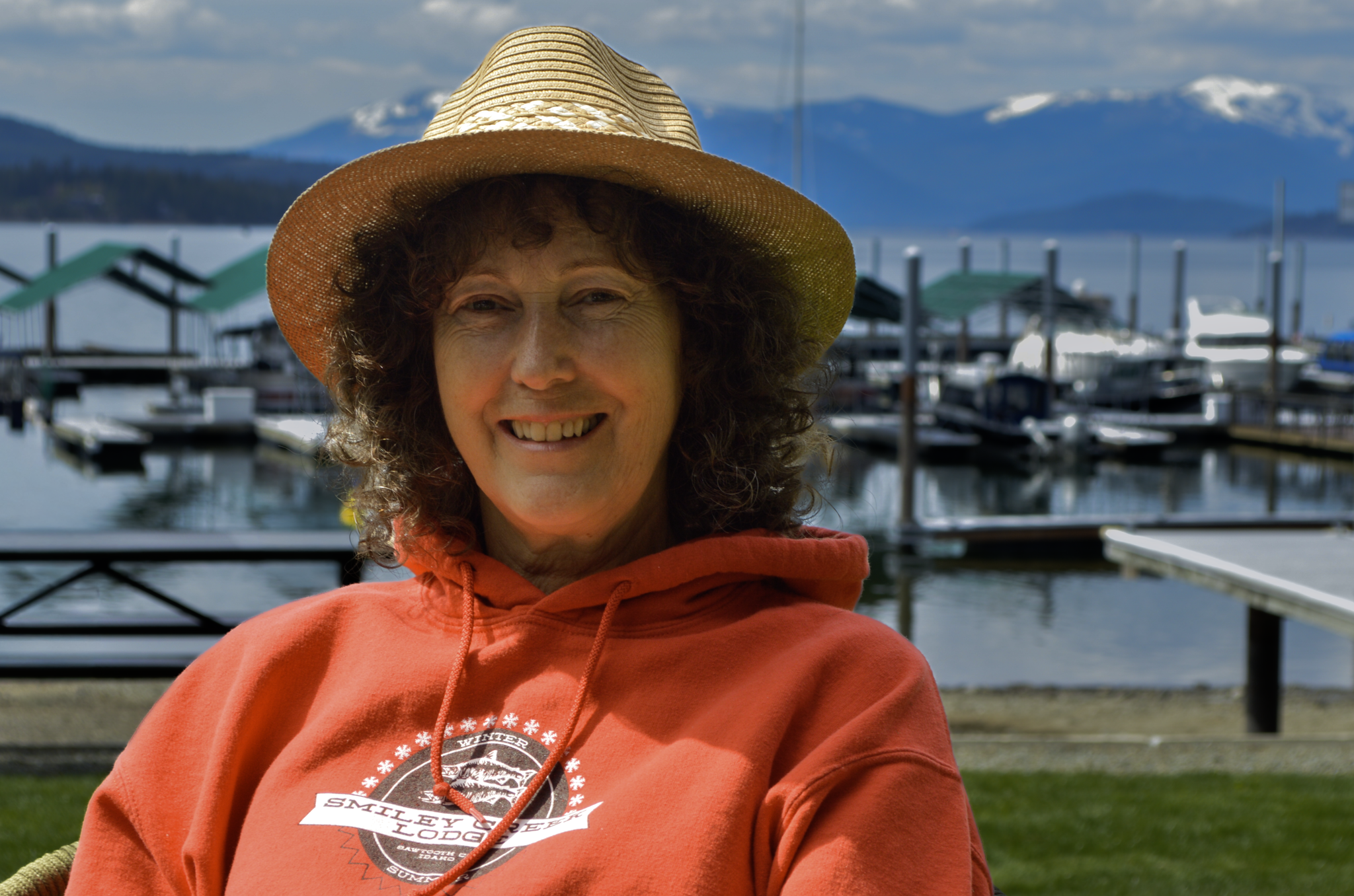 A woman with curly hair wearing a straw hat and an orange hoodie, smiling at a marina with boats docked on the water and snow-capped mountains in the background.
