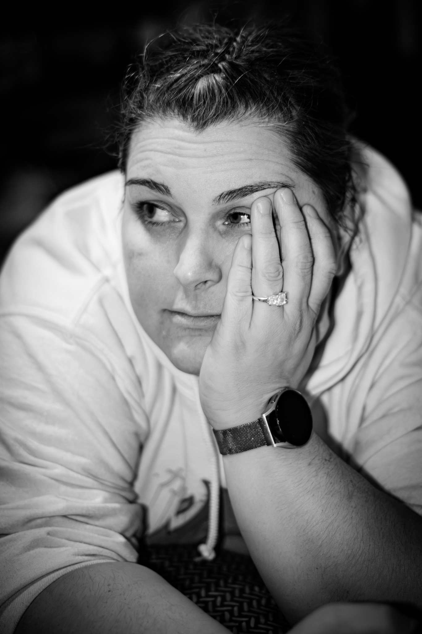 A black and white photo of a woman resting her head on her hand, looking to the side. She is with short hair, wearing a white shirt, a watch, and a ring, sitting in a relaxed pose with her elbows on her knees.