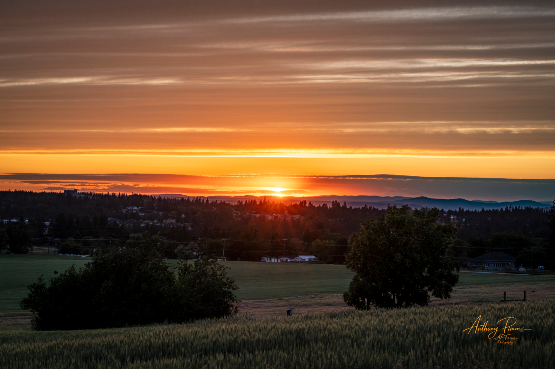 Sunset over a landscape with trees, fields, and distant hills, with colorful clouds in the sky.