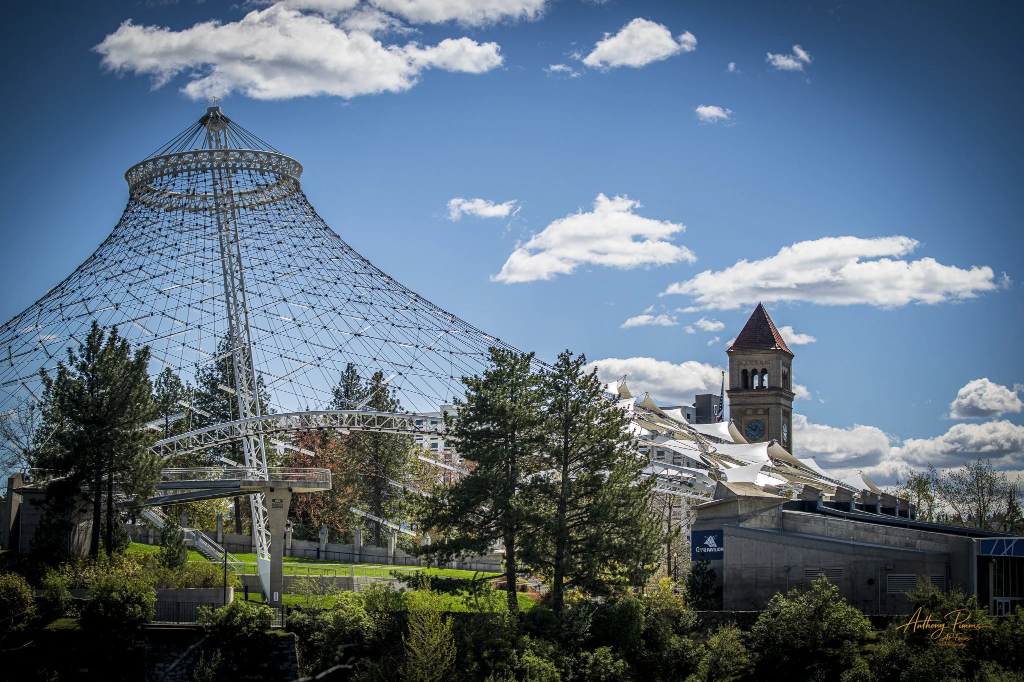 A large amusement park ride with a conical shape made of metal and rope is visible in the foreground, with trees below. In the background, there is a stone clock tower with a pointed roof, and a modern structure with a white, tent-like roof. The sky 