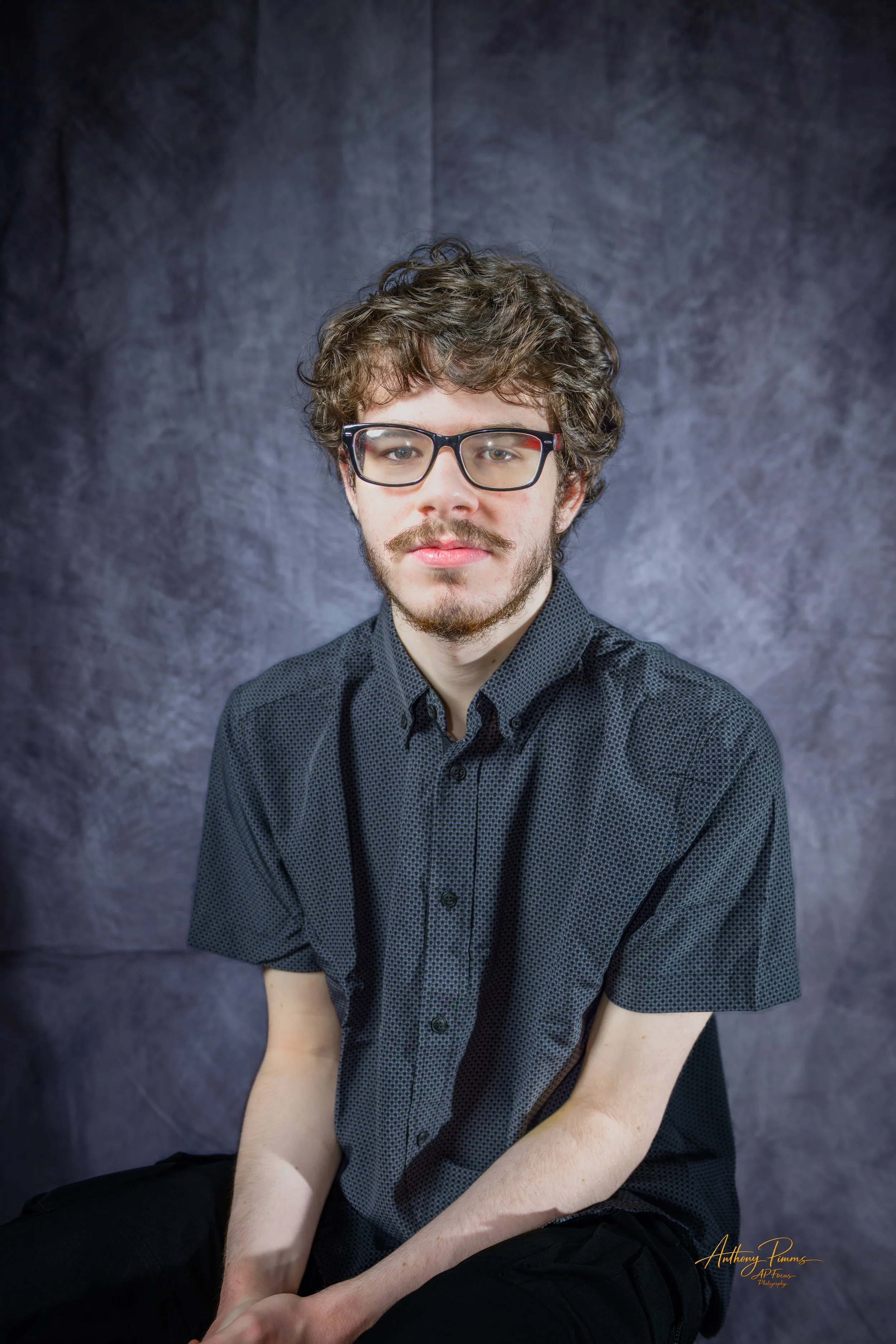 Portrait of a young man with curly brown hair, glasses, and a slight beard wearing a dark button-up shirt, sitting against a dark mottled background.