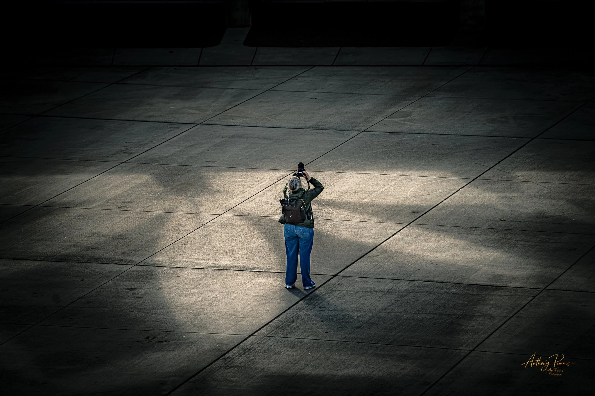 A woman with gray hair wearing a dark jacket and blue jeans is standing on a large tiled surface, taking a photo with a camera, illuminated by a soft light creating a spotlight effect.