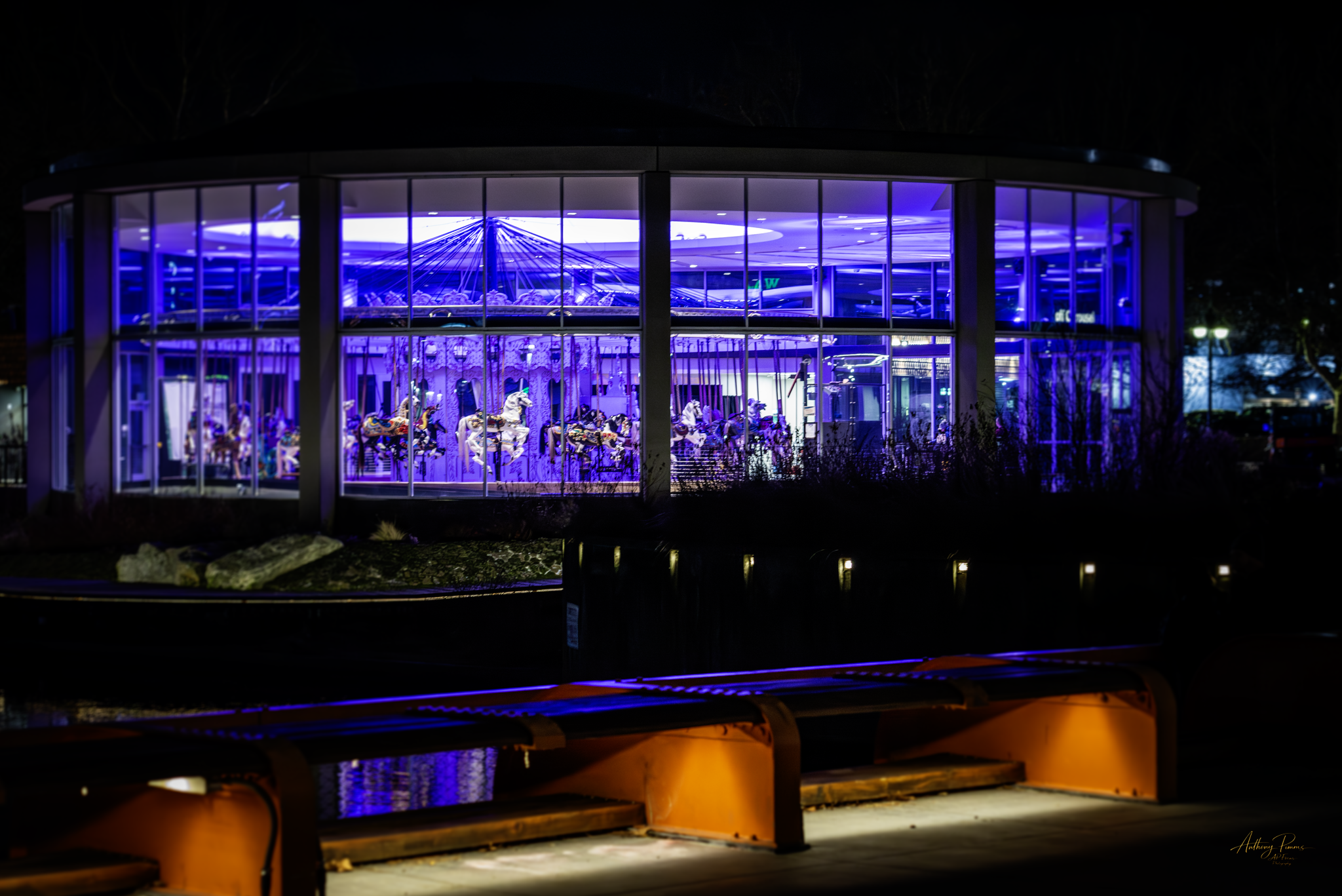 Night view of a carousel inside a glass building illuminated with blue lights, visible through the glass panels.