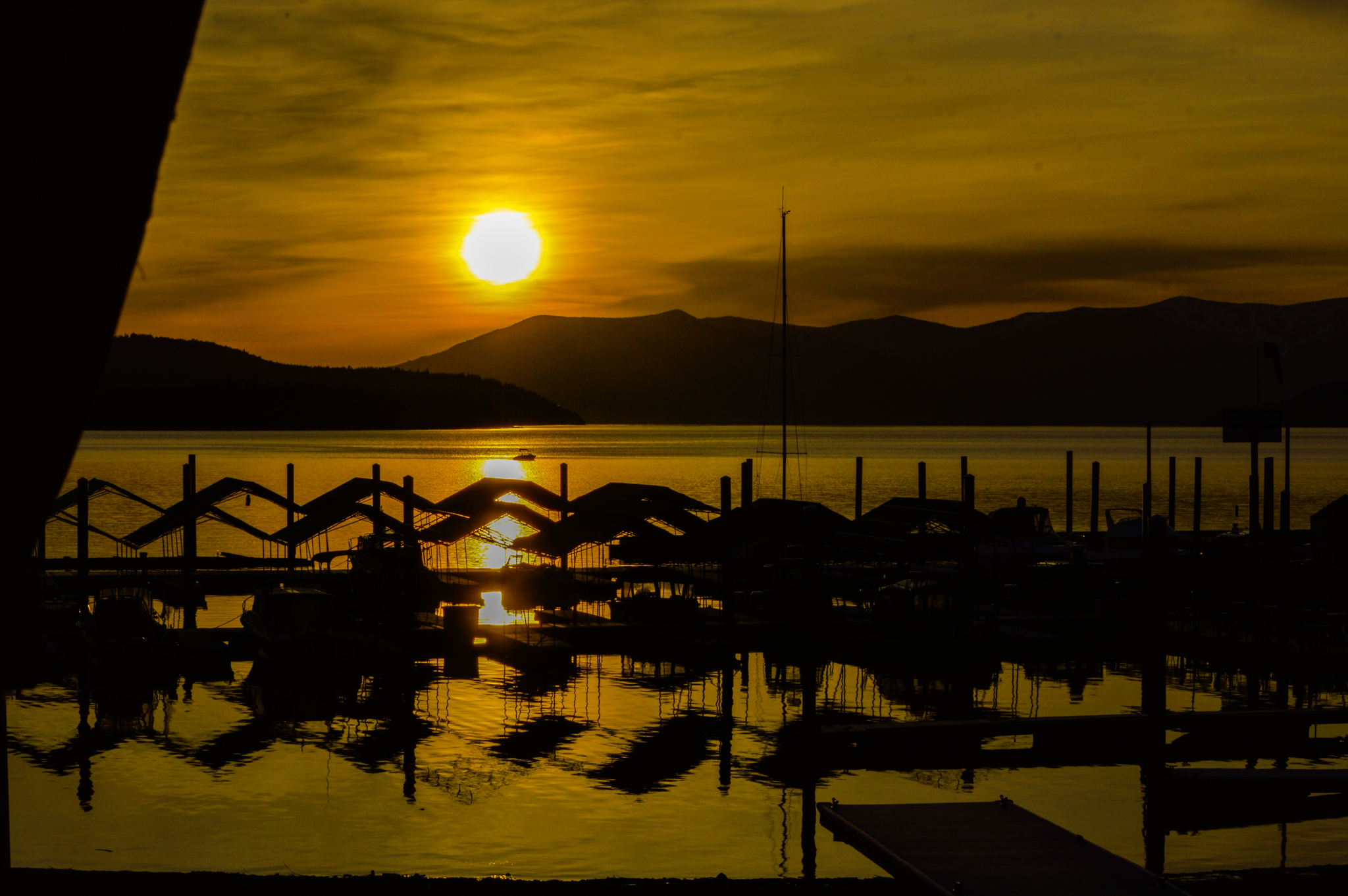 Sunset over a body of water with mountains in the background and boats near a dock, with silhouette umbrellas in the foreground.
