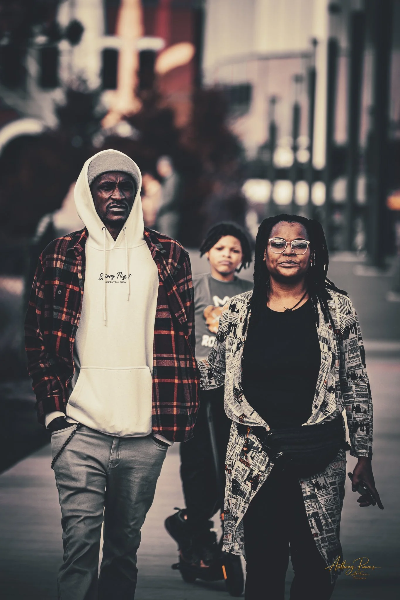 A group of three diverse people walking on a city sidewalk, with buildings in the background. The person in front is wears glasses and a newspaper-patterned coat, the person to their left has dreadlocks and a gray shirt, and the person to their right