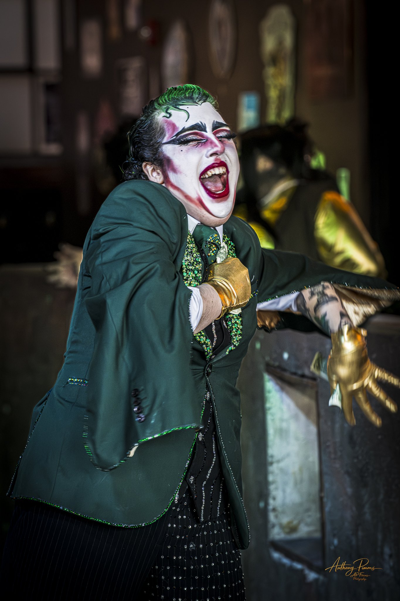 A person dressed as a clown with white face paint, colorful makeup, and a green and black costume, appears to be shouting or singing with a joyful expression.