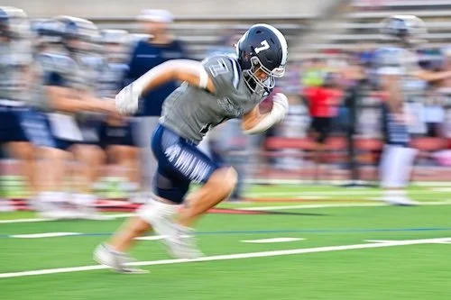 A football player in a gray shirt and blue shorts running on a field with a helmet on, holding a football, with other players and spectators blurred in the background.