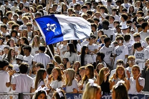 A large group of students wearing white shirts gathered outdoors, with some holding a white and blue flag.