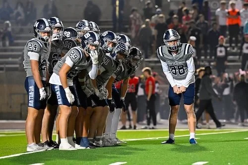 Group of football players in black and white uniforms lined up on the field, preparing for a play, with spectators in the background.