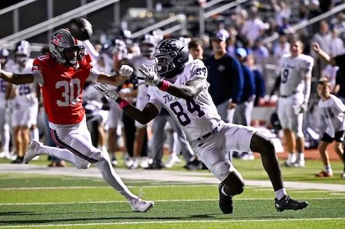 A football player in a red jersey with the number 30 running while being tackled by a football player in a white jersey with the number 84 on a football field during a game.