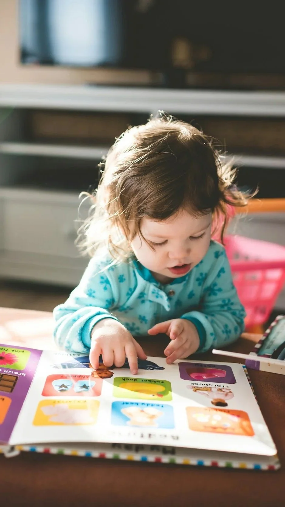 A young girl with brown hair wearing a light blue pajamas with darker blue flowers, sitting at a table and pointing to a colorful children's book with pictures and words.