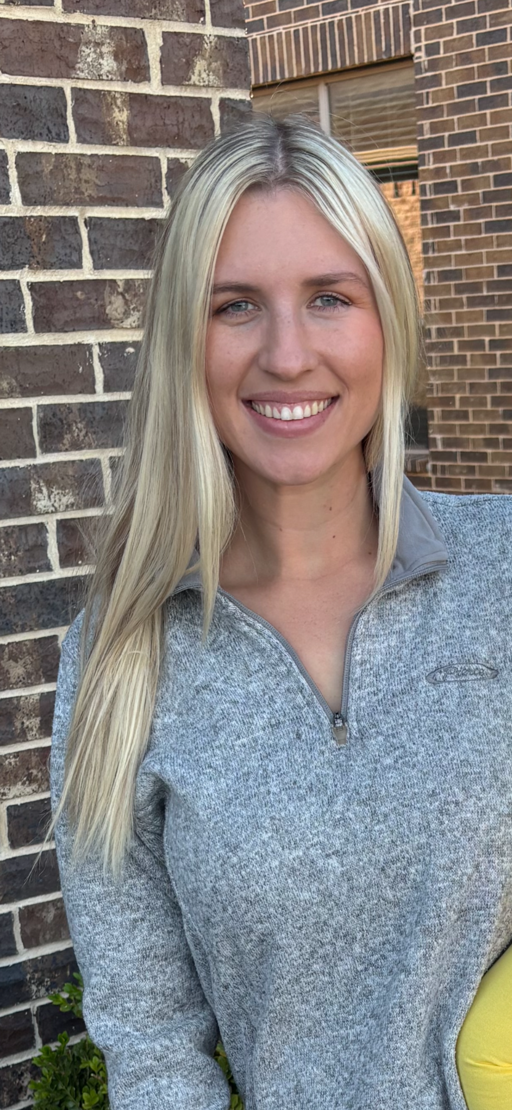 A young woman with long blonde hair, fair skin, and blue eyes smiling, standing outdoors in front of a brick wall.