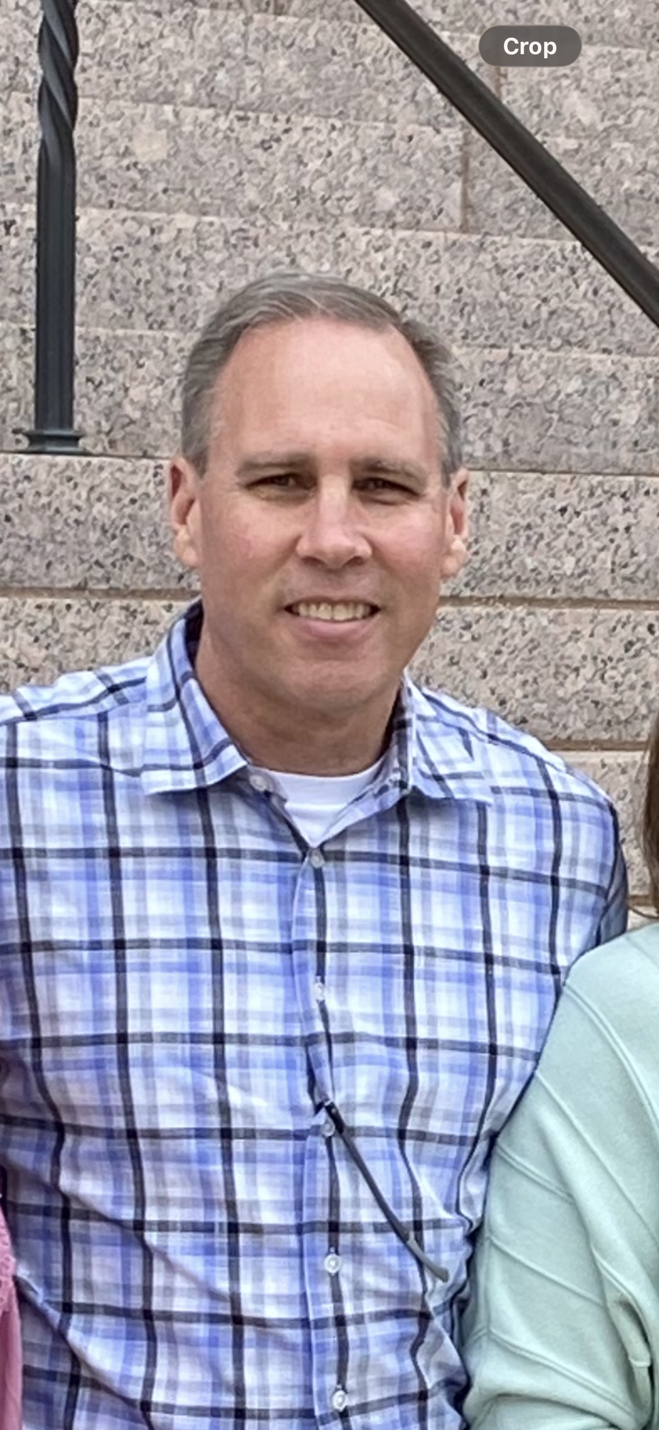 A man with short, light brown hair, wearing a blue and white plaid button-down shirt, smiling while standing in front of a stone wall with a staircase.