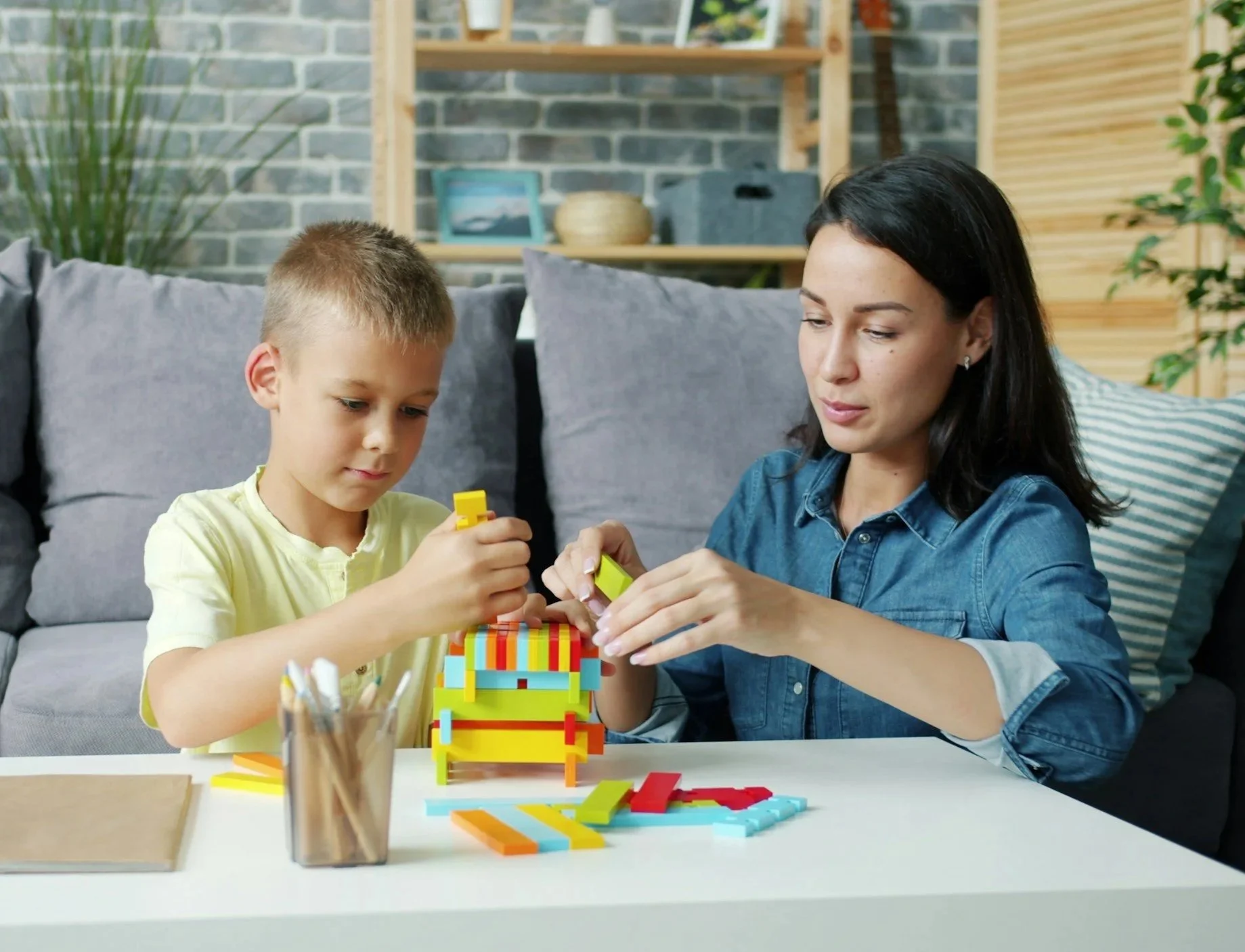 A woman and a young boy playing with colorful building blocks at a white table in a living room with a gray couch, green plants, and wooden shelves in the background.