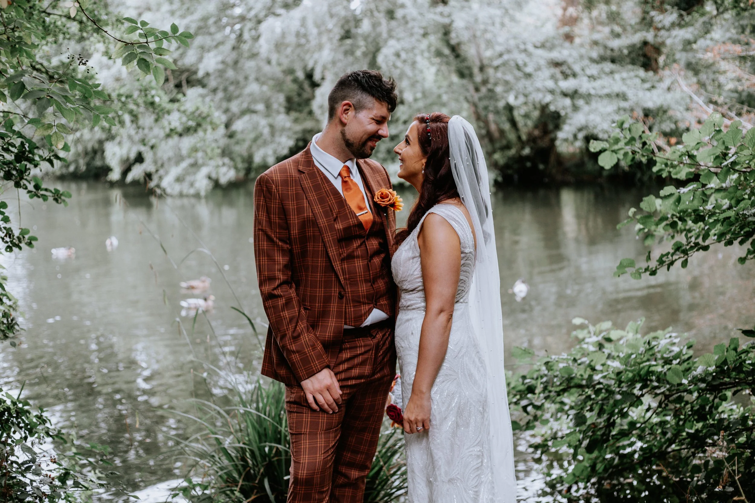 A bride and groom stand close together by a lake surrounded by trees, looking at each other lovingly during their wedding.