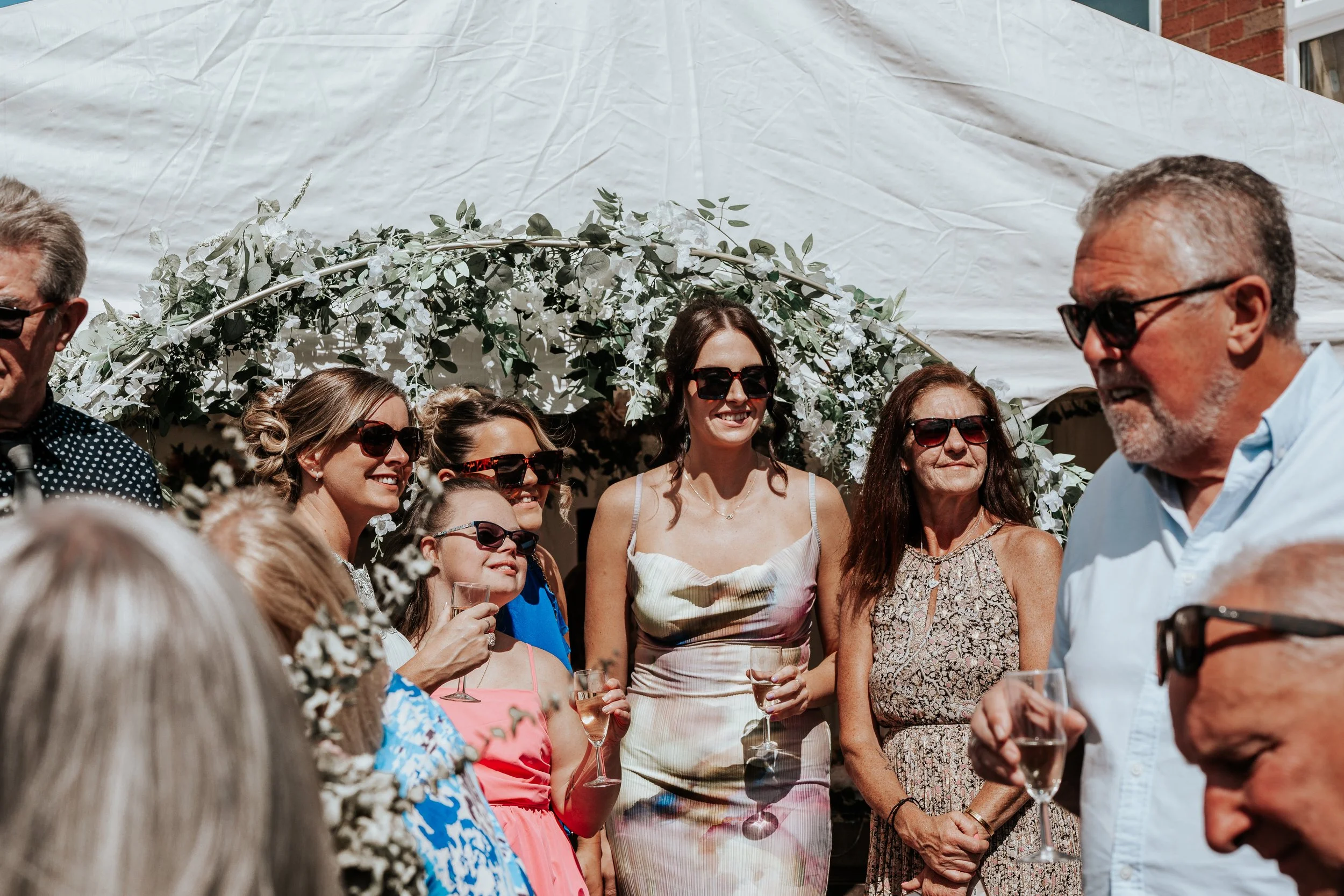 A group of women and a man at a wedding celebration, many holding glasses of champagne, standing under a white floral arch, all wearing sunglasses.