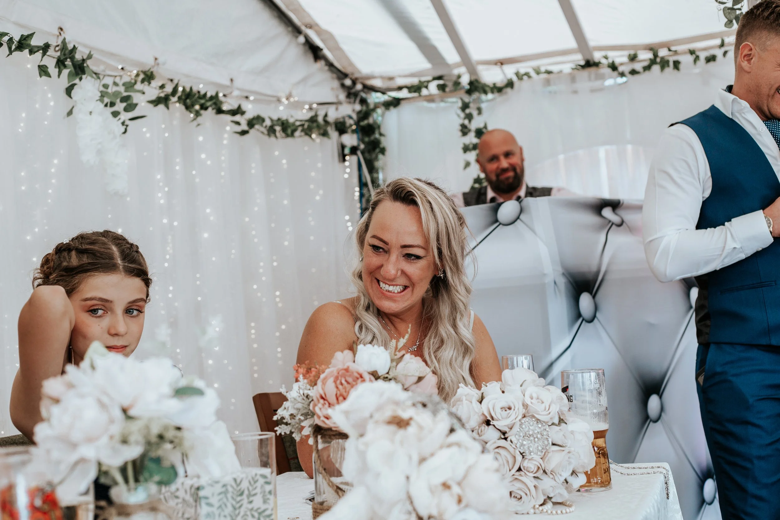 A woman with blonde hair smiling at a wedding reception, surrounded by floral arrangements and wedding decorations, with a young girl sitting nearby and a man in a vest seen in the background.