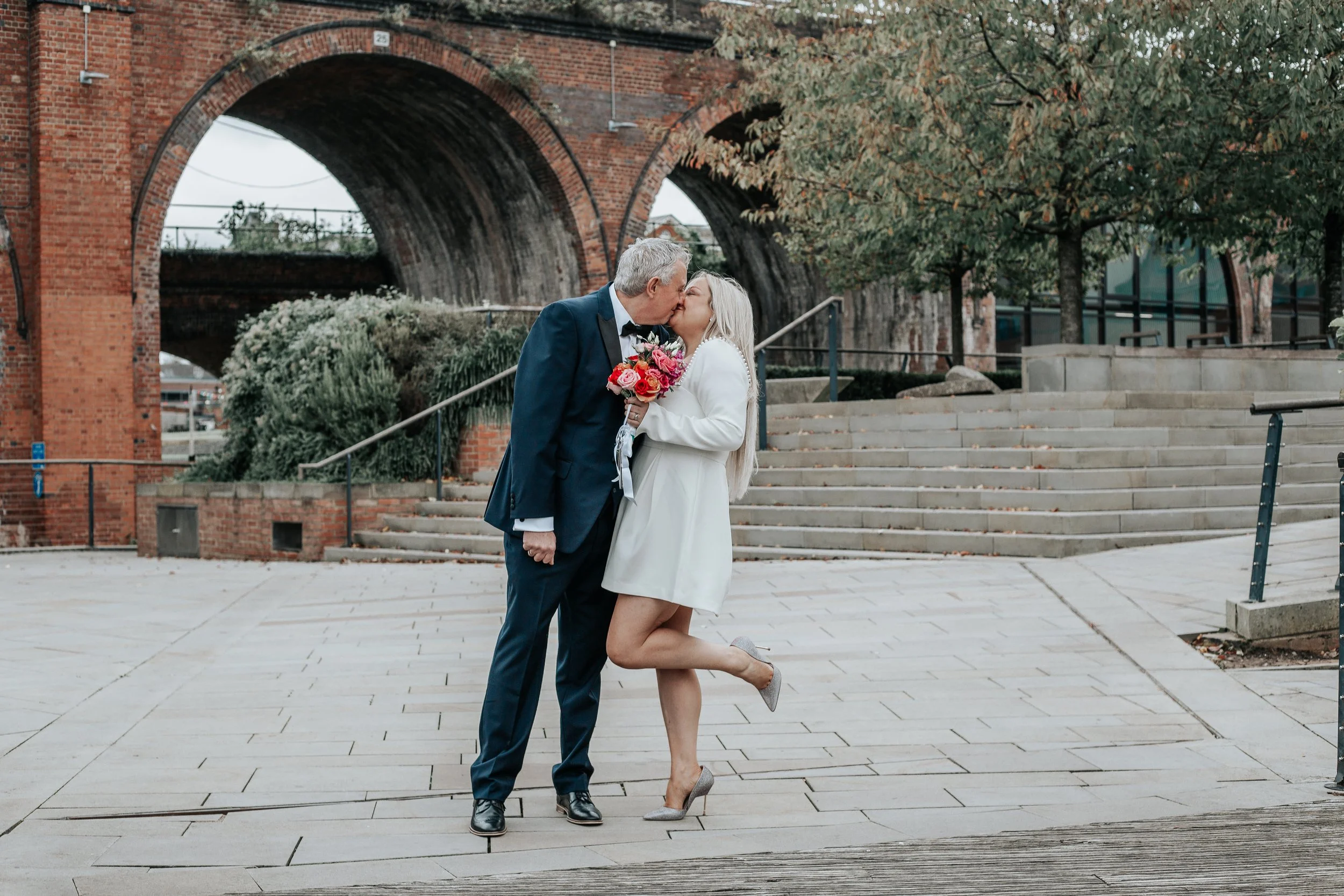 A couple sharing a kiss in a public outdoor space with brick arches and steps, with the woman holding a bouquet of flowers.