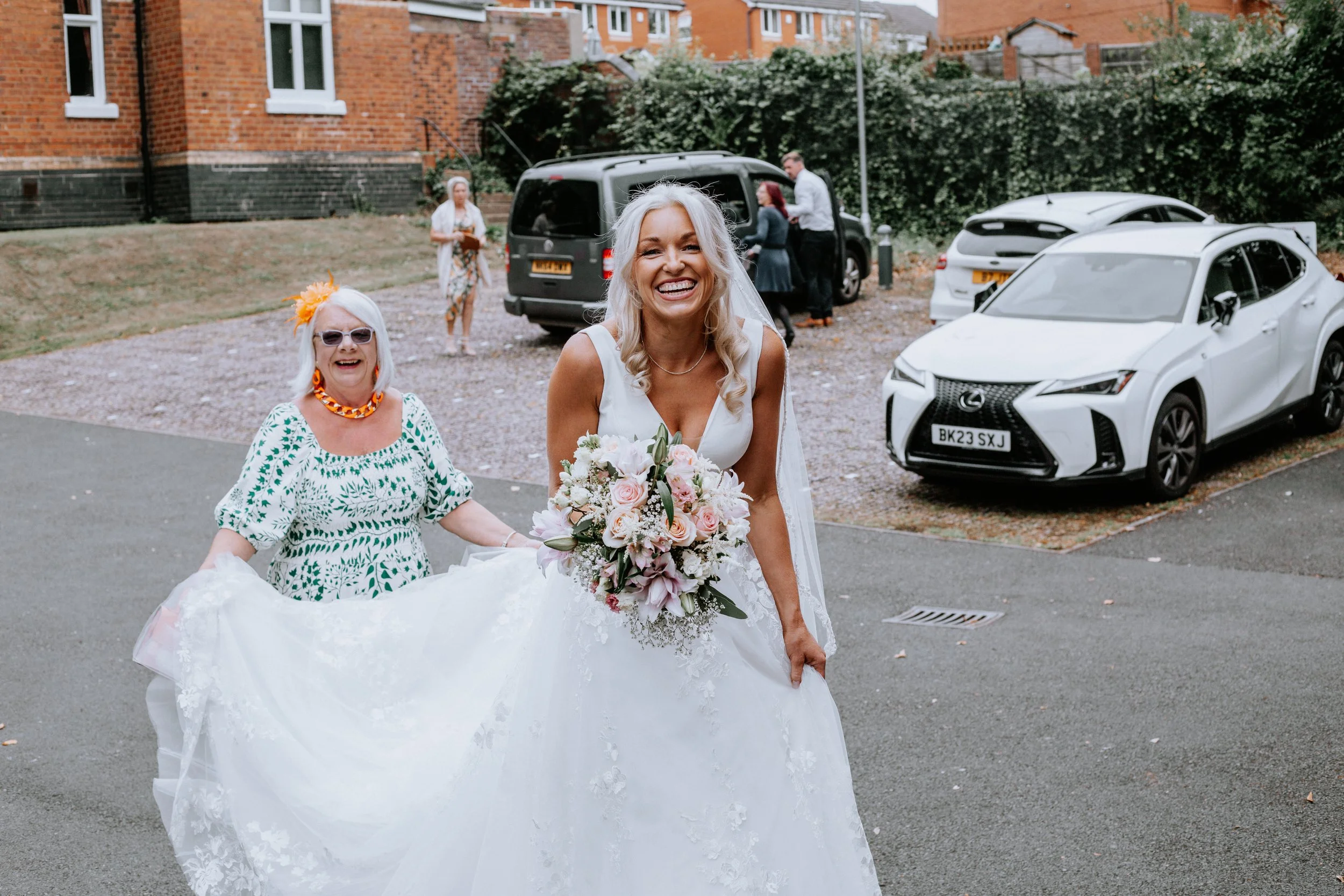 A bride in a white wedding dress holding a bouquet of pink and white flowers, smiling, walking outdoors. An older woman in sunglasses and a white dress with green patterns with an orange necklace and flower headband is beside her, also smiling. In the background, several people are near a black van and a white car, with a brick building and greenery behind them.
