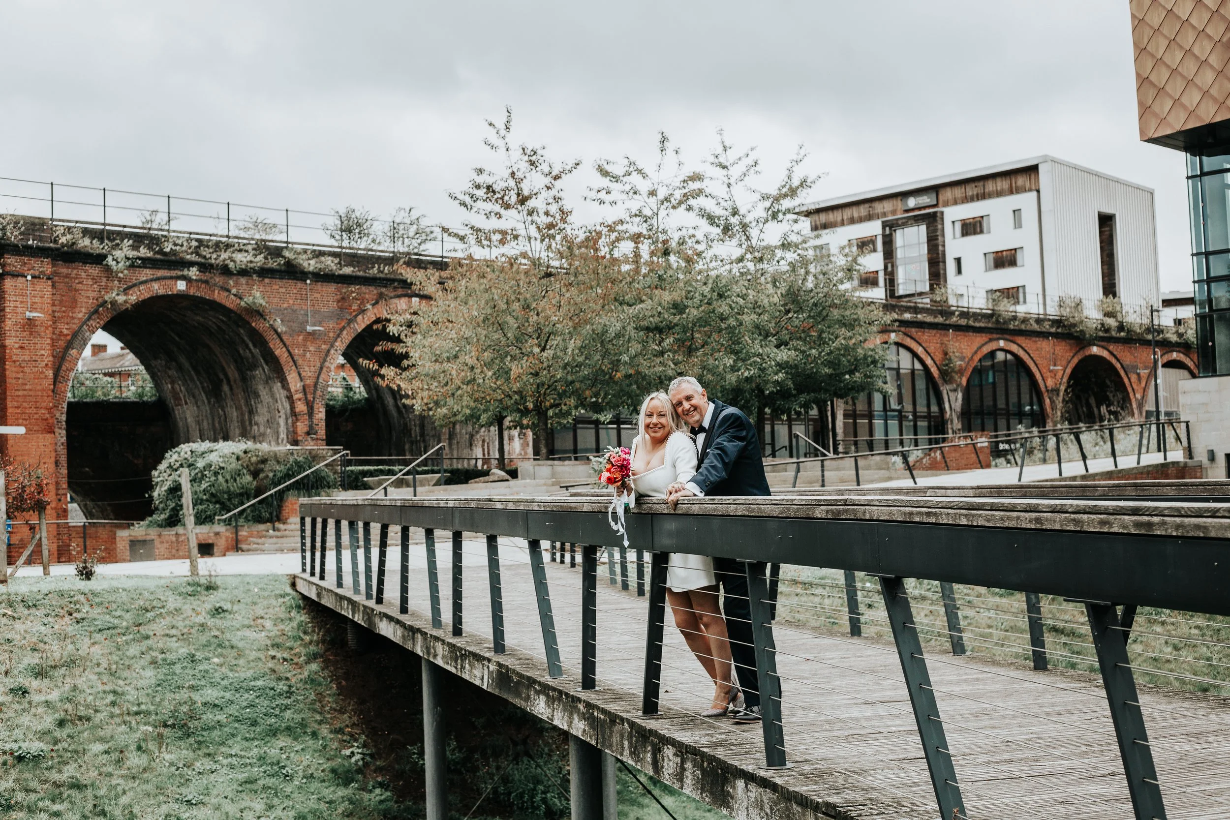 A newlywed couple in wedding attire, smiling and embracing on a wooden bridge in an urban park with brick arches and modern buildings in the background.
