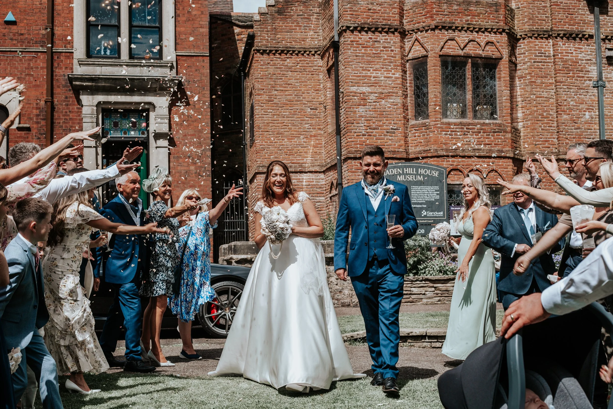 Bride and groom smiling as they walk through a crowd of wedding guests outside a brick building, celebrating their wedding day.