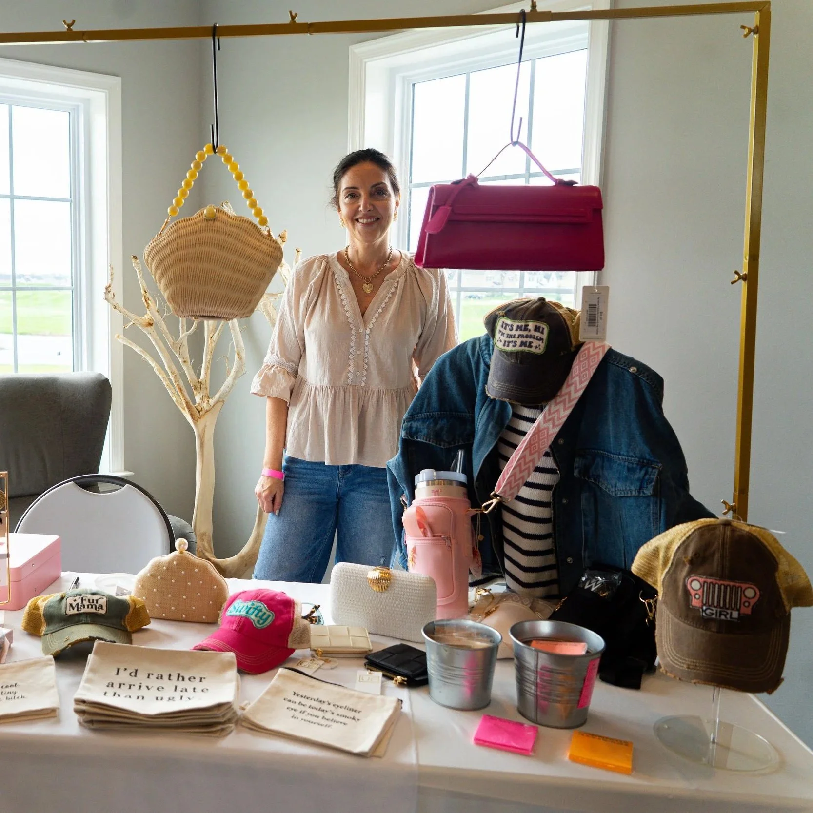 A woman smiling standing behind a table filled with various hats, accessories, and small items at a craft or flea market inside a room with large windows.