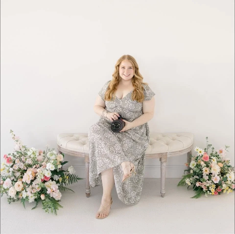 A woman with long, wavy red hair sitting on a beige upholstered bench, holding a camera, flanked by floral arrangements, in a minimalist room with a white wall background.
