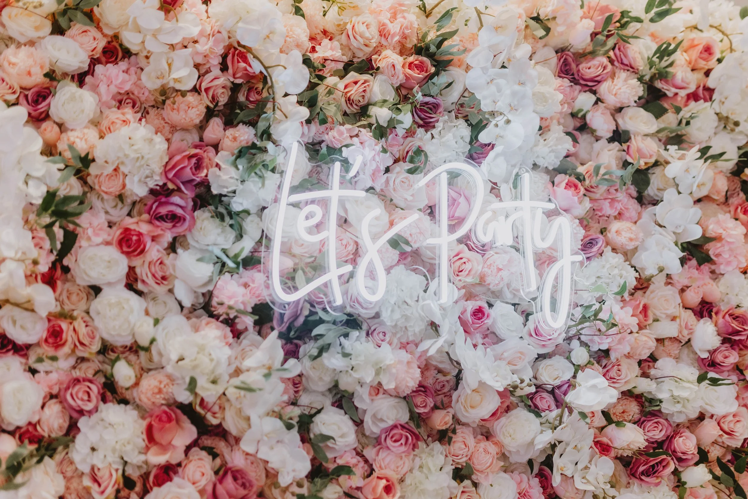 A wall of pink, white, and cream roses and white orchids with a neon sign that says 'let's party'.