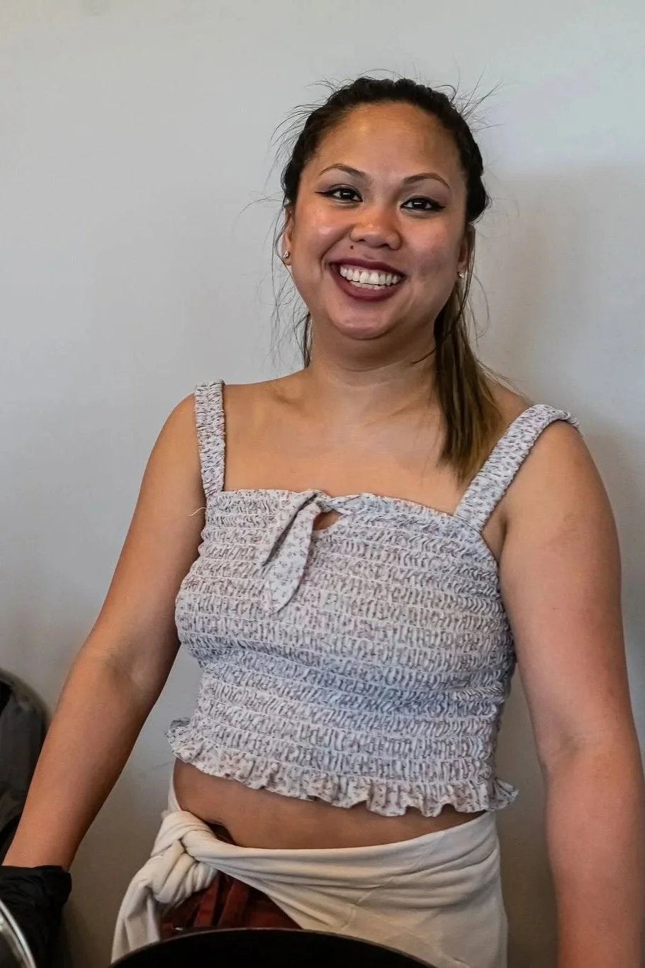 A smiling woman with brown hair tied back, wearing a sleeveless, patterned crop top and light-colored pants, standing against a plain wall.